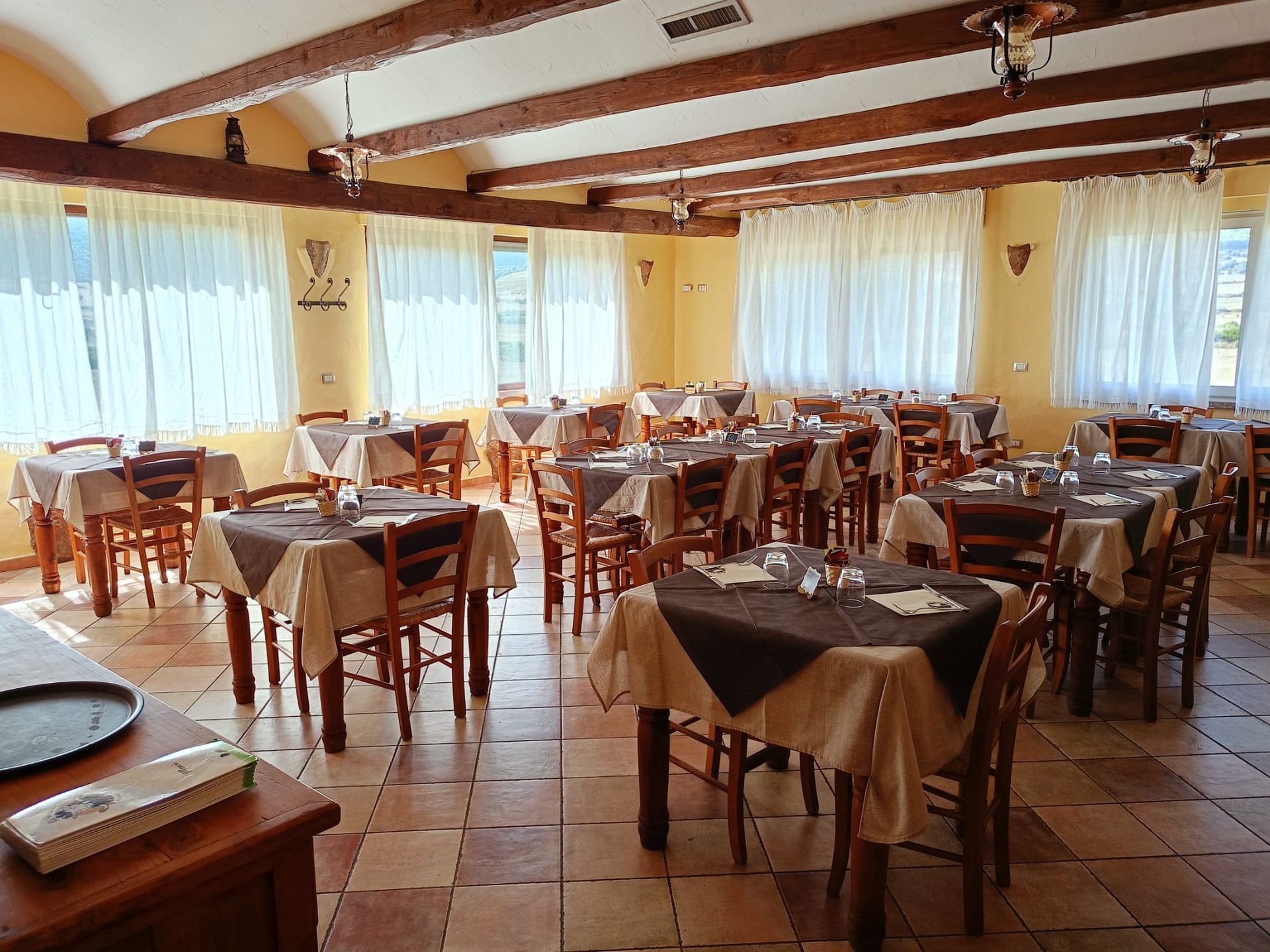 Sala da pranzo vuota di un ristorante con tavoli in legno, pavimento a scacchi e grandi finestre luminose.