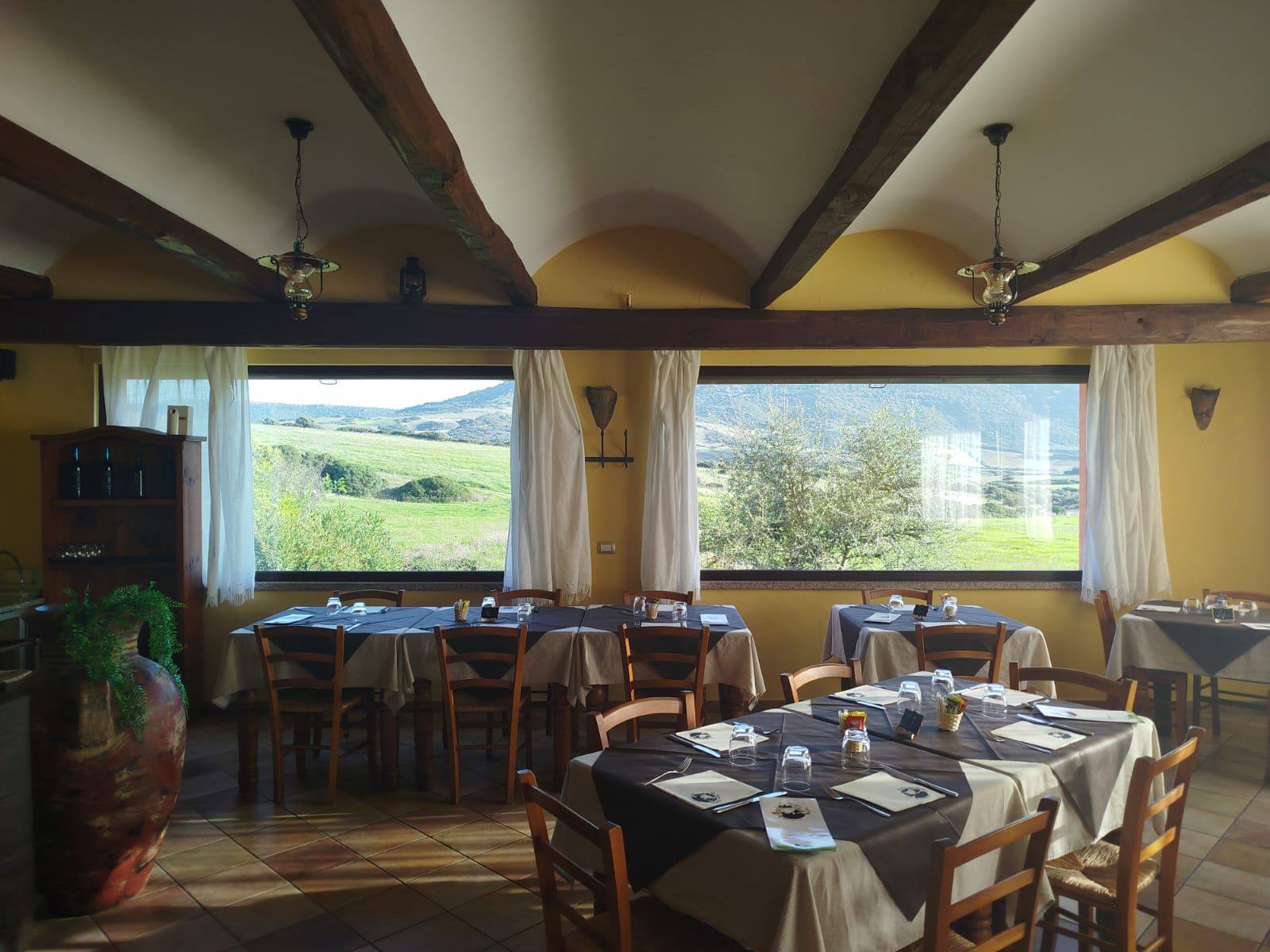 Accogliente sala da pranzo con travi in ​​legno, tavoli apparecchiati e ampie finestre che si affacciano su un paesaggio verdeggiante.