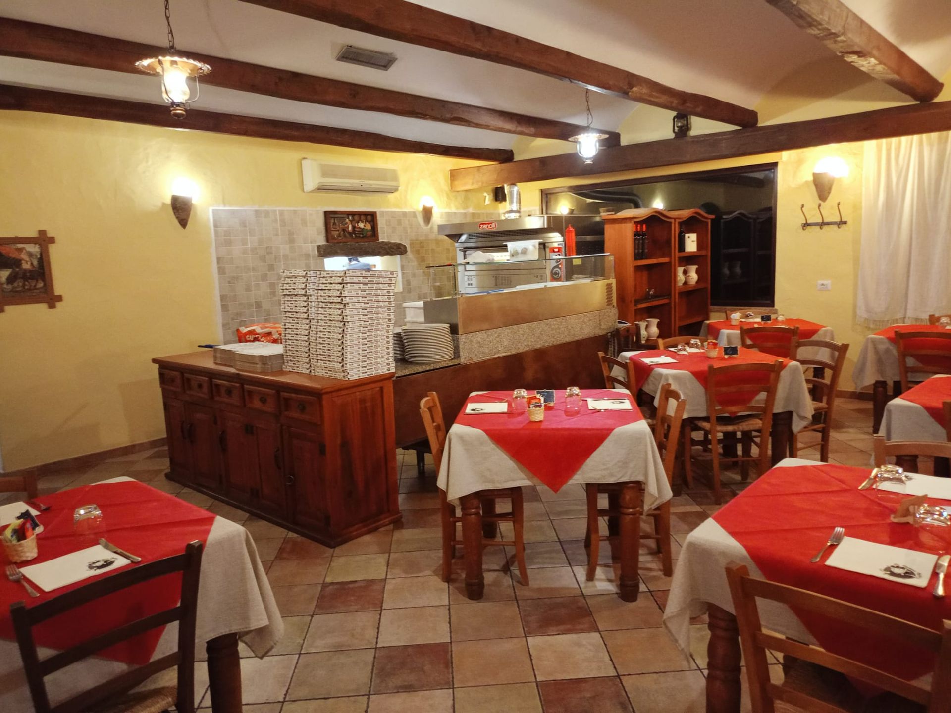 Sala da pranzo vuota di un ristorante con tovaglie rosse, travi in ​​legno e pavimento piastrellato.