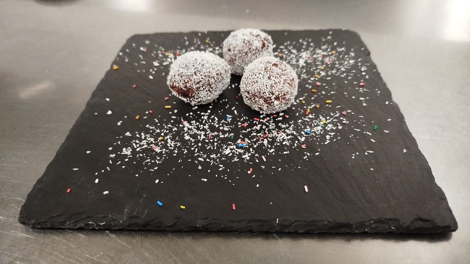 Quattro tartufi da dessert ricoperti di cocco, disposti su un piatto di ardesia nera e cosparsi di zuccherini colorati.
