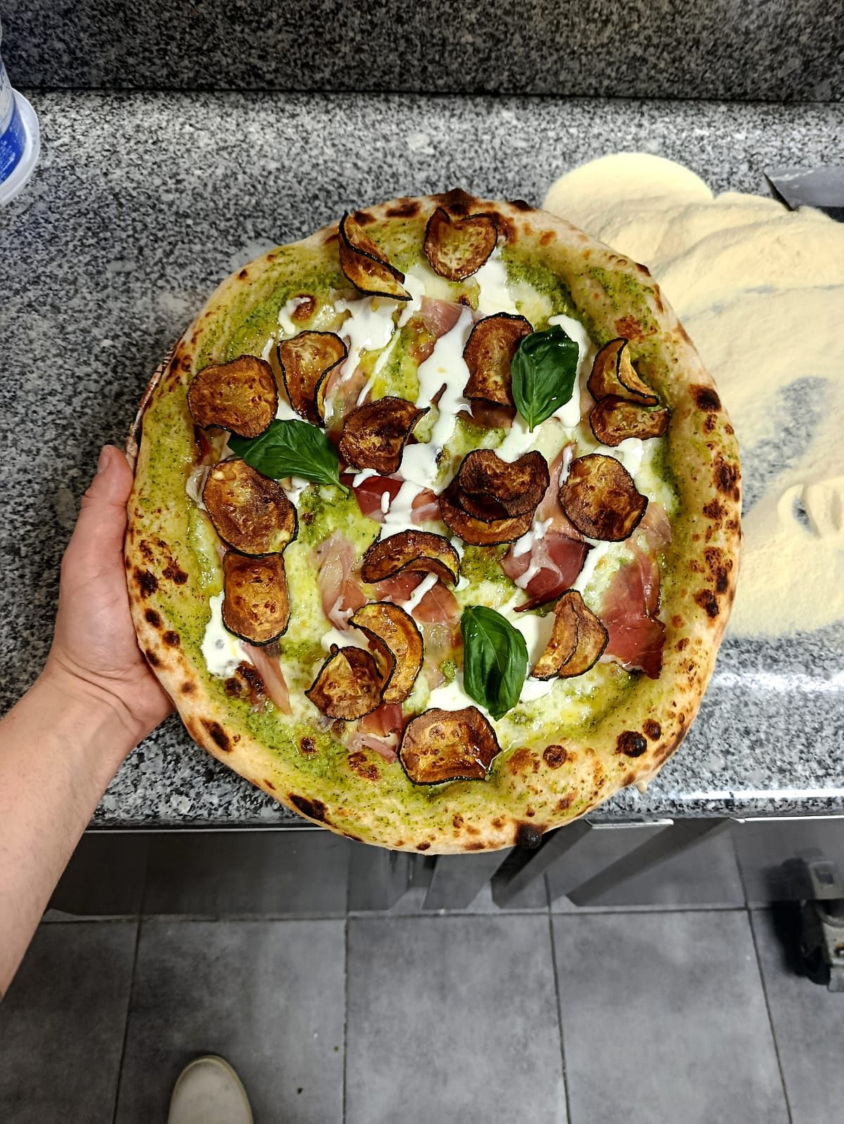 Una mano regge una pizza condita con pancetta, peperoni verdi e formaggio, appoggiata su un bancone grigio con un piatto bianco.