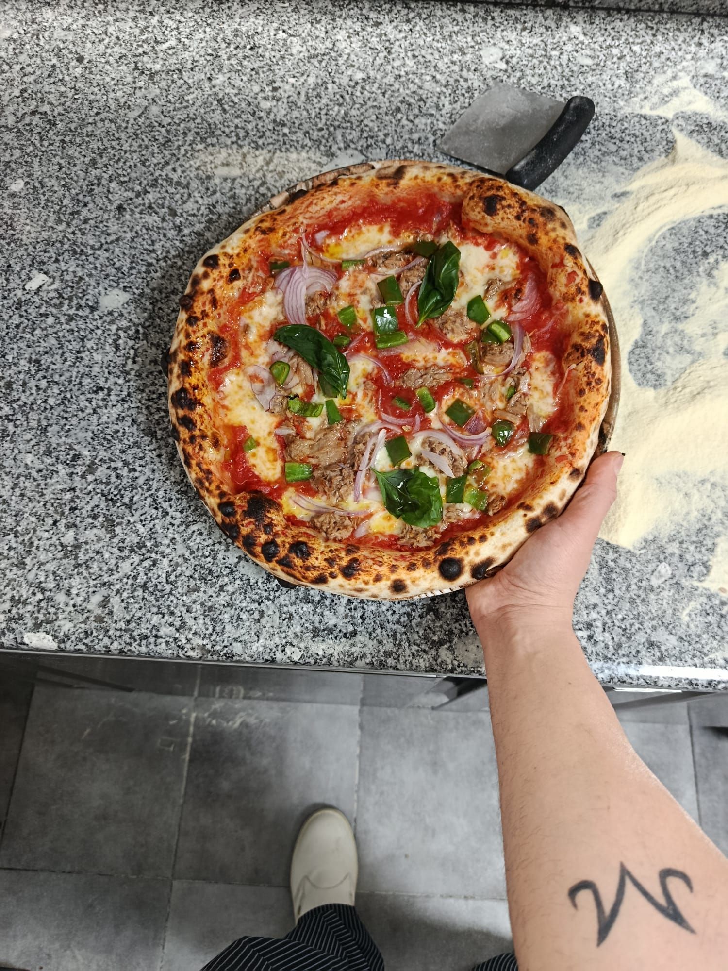 Una mano che regge una pizza appena sfornata su un piano di lavoro in granito, con una pala per pizza sullo sfondo.