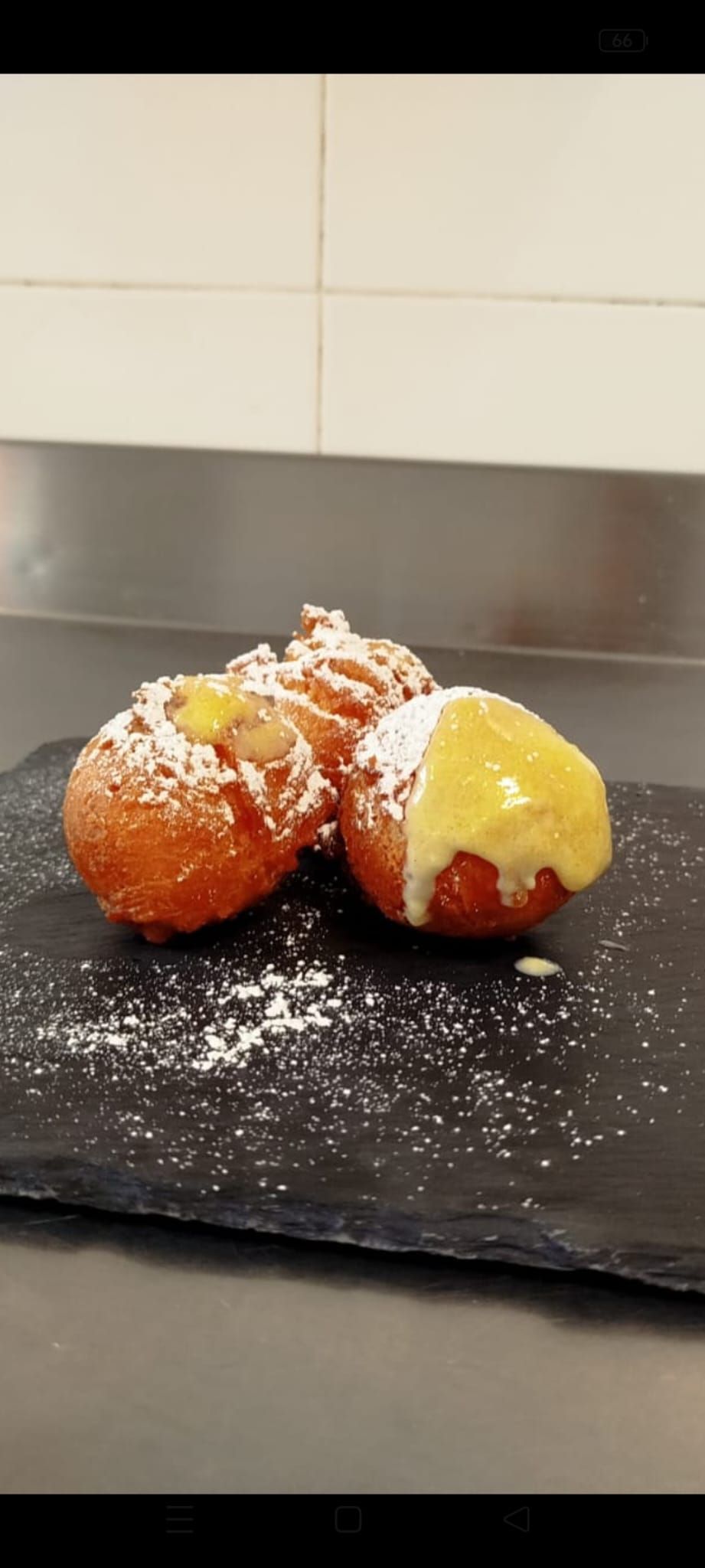 Tre palline di pasta fritta cosparse di zucchero a velo, servite con panna e zucchero a velo su un piatto nero.