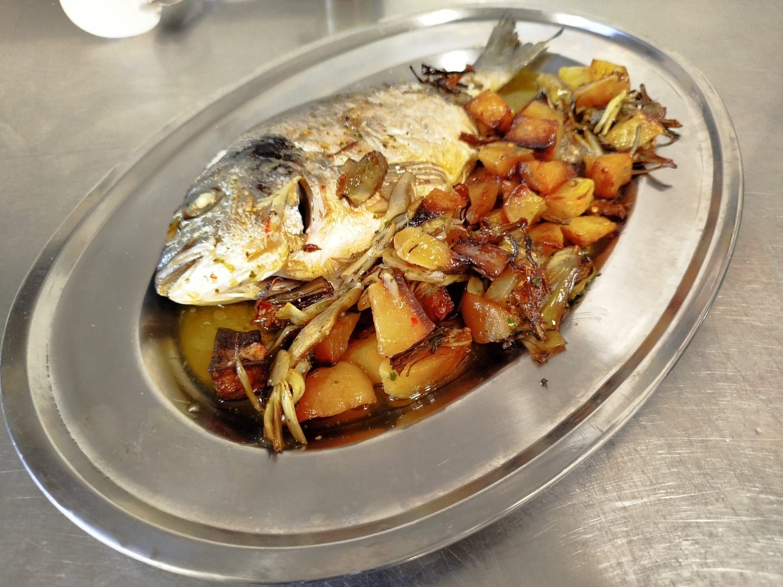 Pesce intero cotto in salsa bruna con verdure a dadini su un vassoio di metallo.