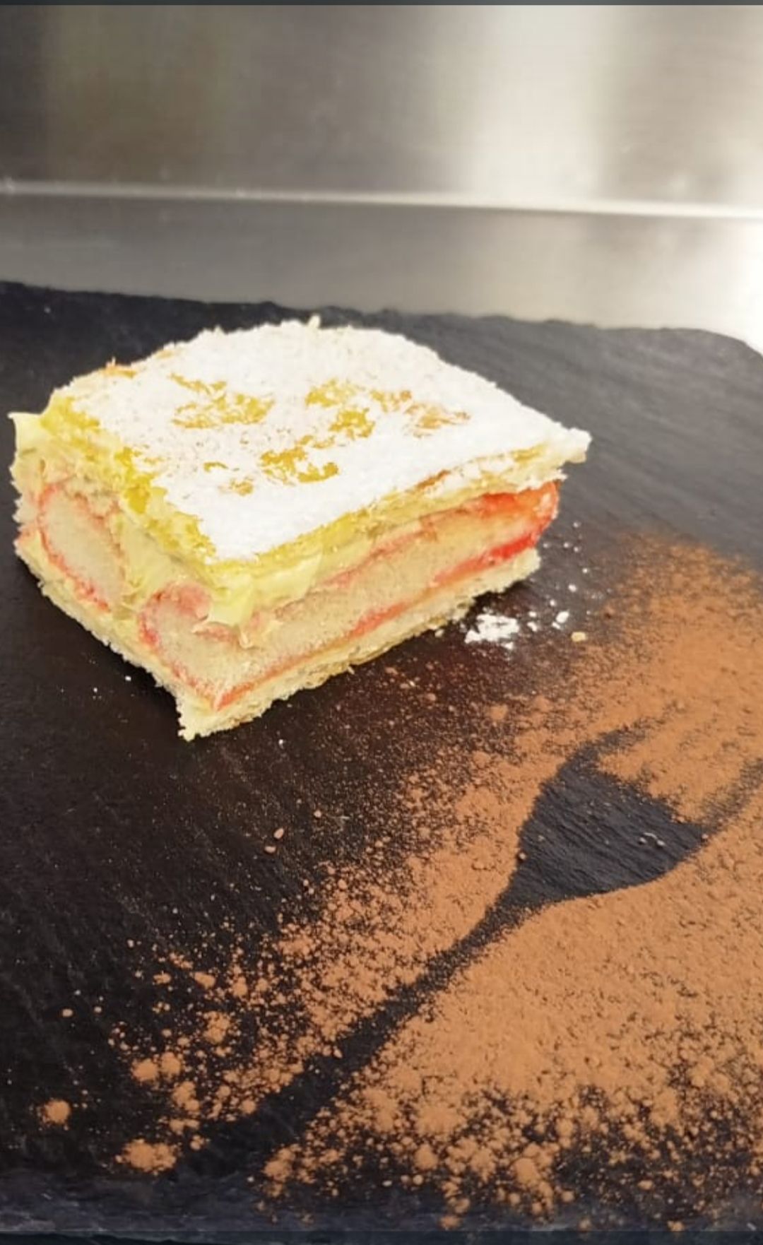Fetta di dessert a strati con crema e ripieno di frutta, spolverata di zucchero a velo su un piatto nero con cacao in polvere.