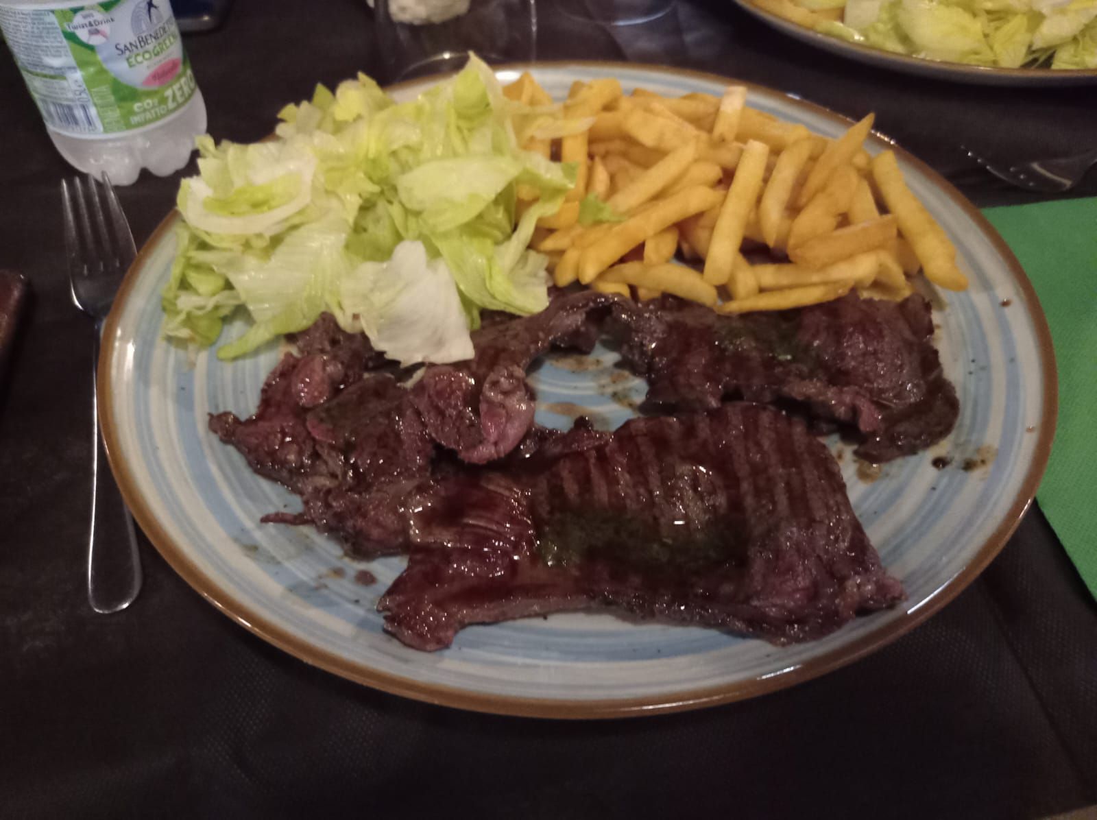 Un piatto con bistecca alla griglia, patatine fritte e insalata di lattuga su un tavolo accanto a una bottiglia d'acqua.