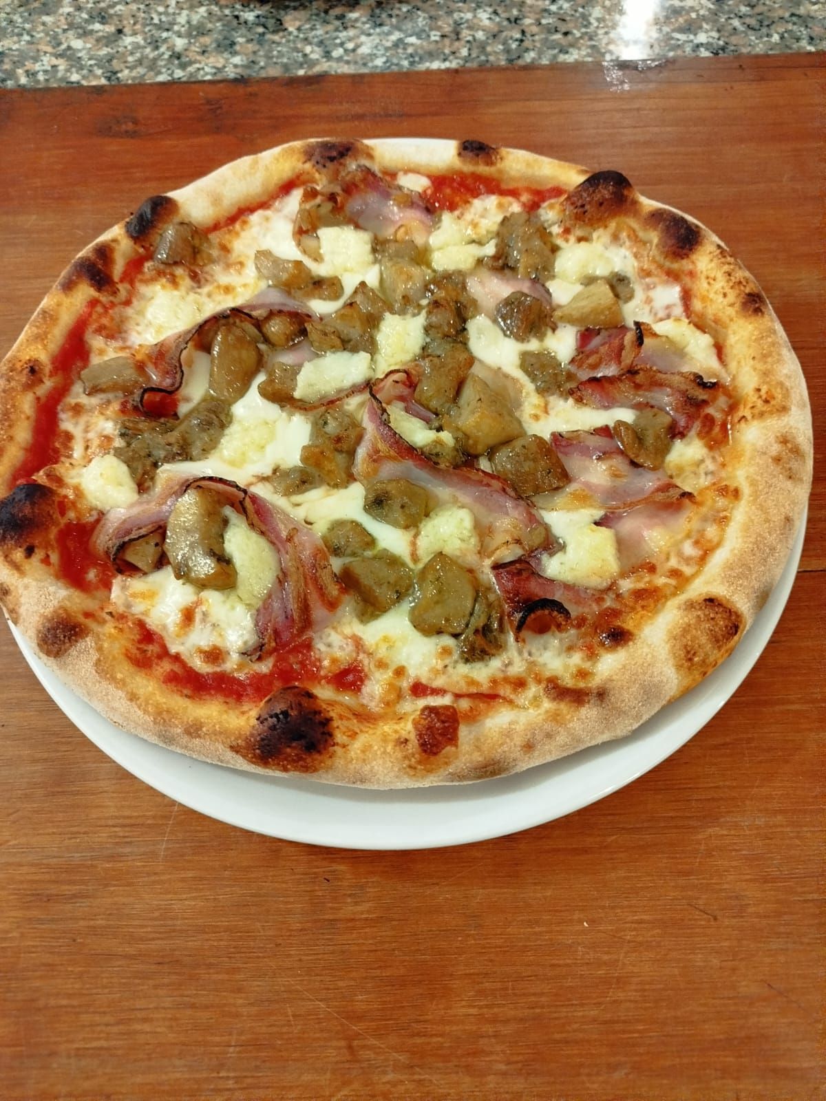 Una pizza con formaggio fuso, funghi e altri condimenti su un piatto bianco appoggiato su un tavolo di legno.