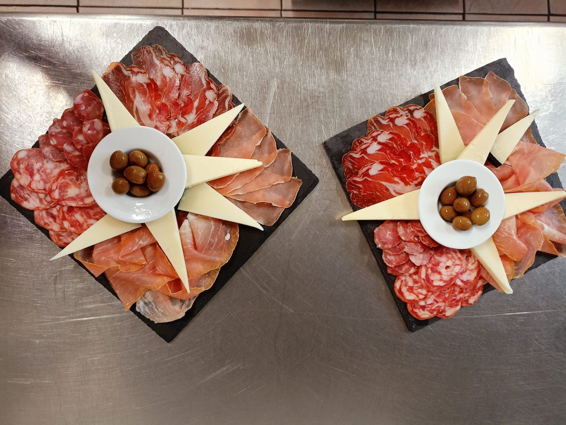 Due taglieri di salumi con affettati, spicchi di formaggio e olive su un tavolo grigio.