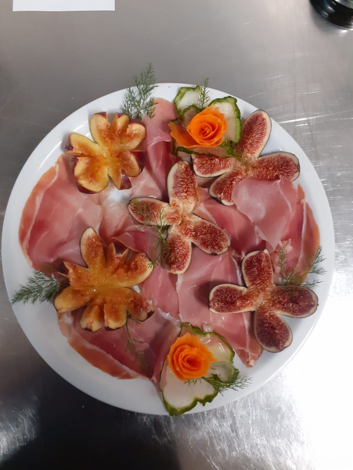 Piatto bianco di salumi, fichi e guarnizioni di fiori d'arancio su un tavolo in acciaio inox.