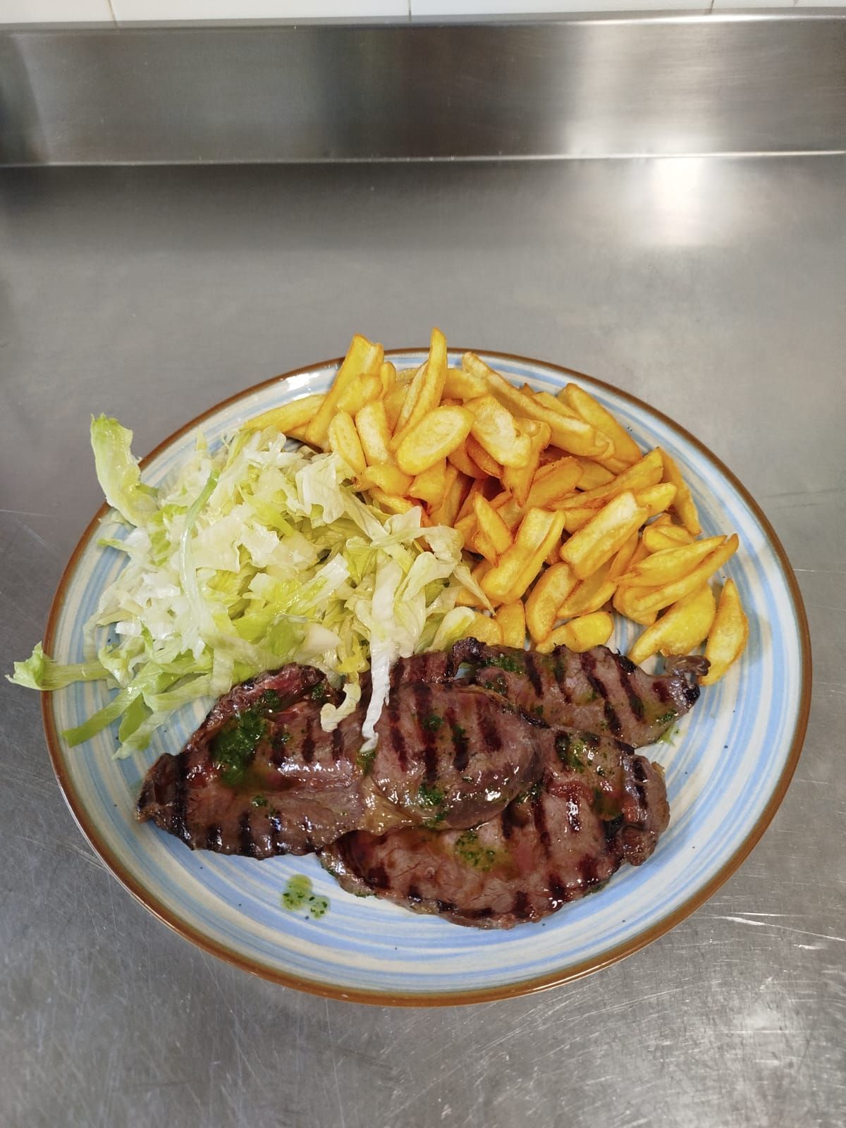 Bistecca alla griglia con patatine fritte e lattuga tritata su un piatto appoggiato su un bancone in acciaio inossidabile.