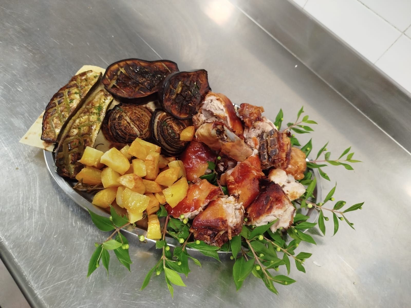 Pollo arrosto, patate e verdure guarnite con erbe aromatiche su un piano di lavoro in metallo.