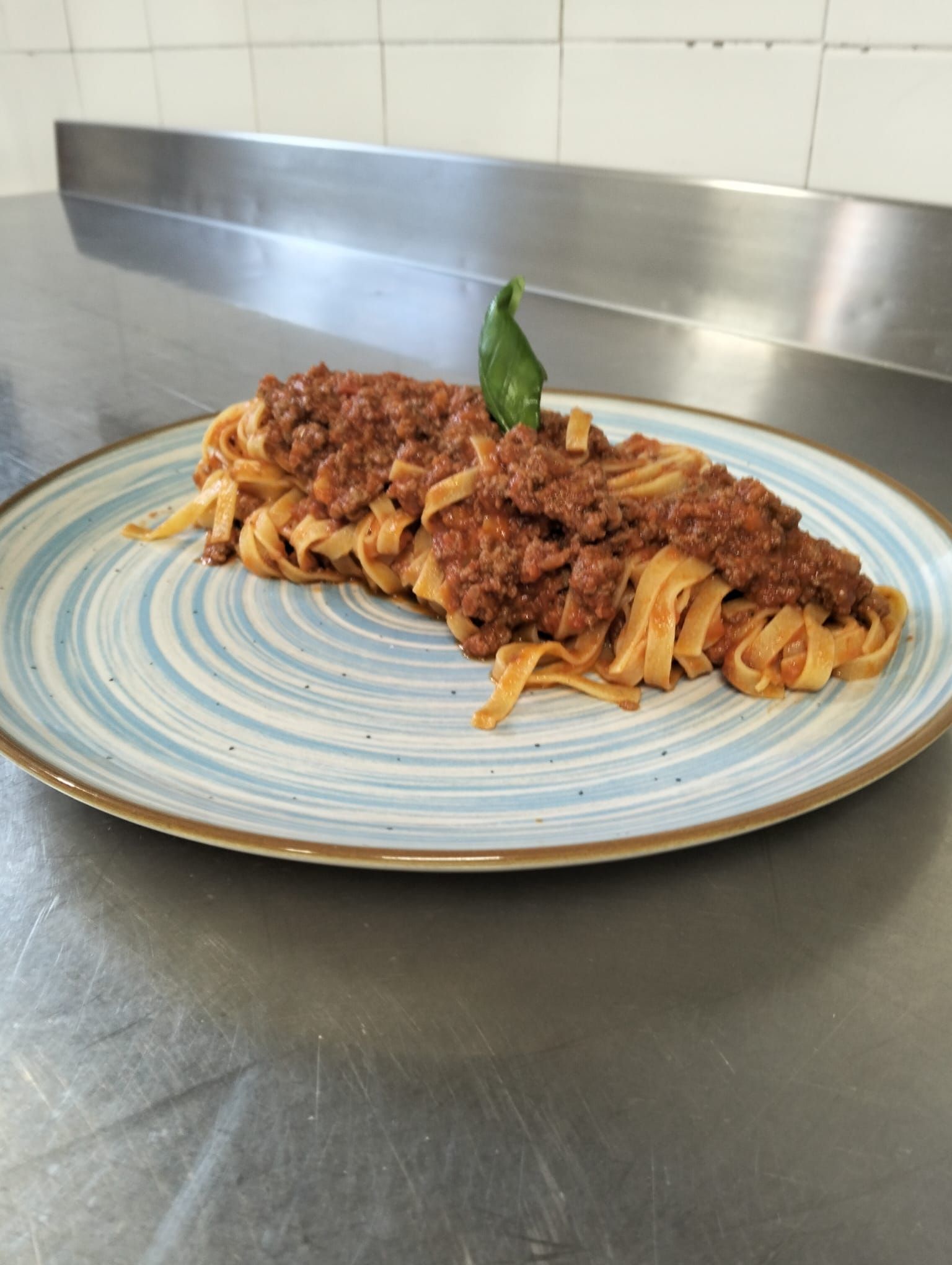 Pasta al ragù su un piatto con bordo blu, guarnita con una foglia di basilico, su un bancone in acciaio inox.