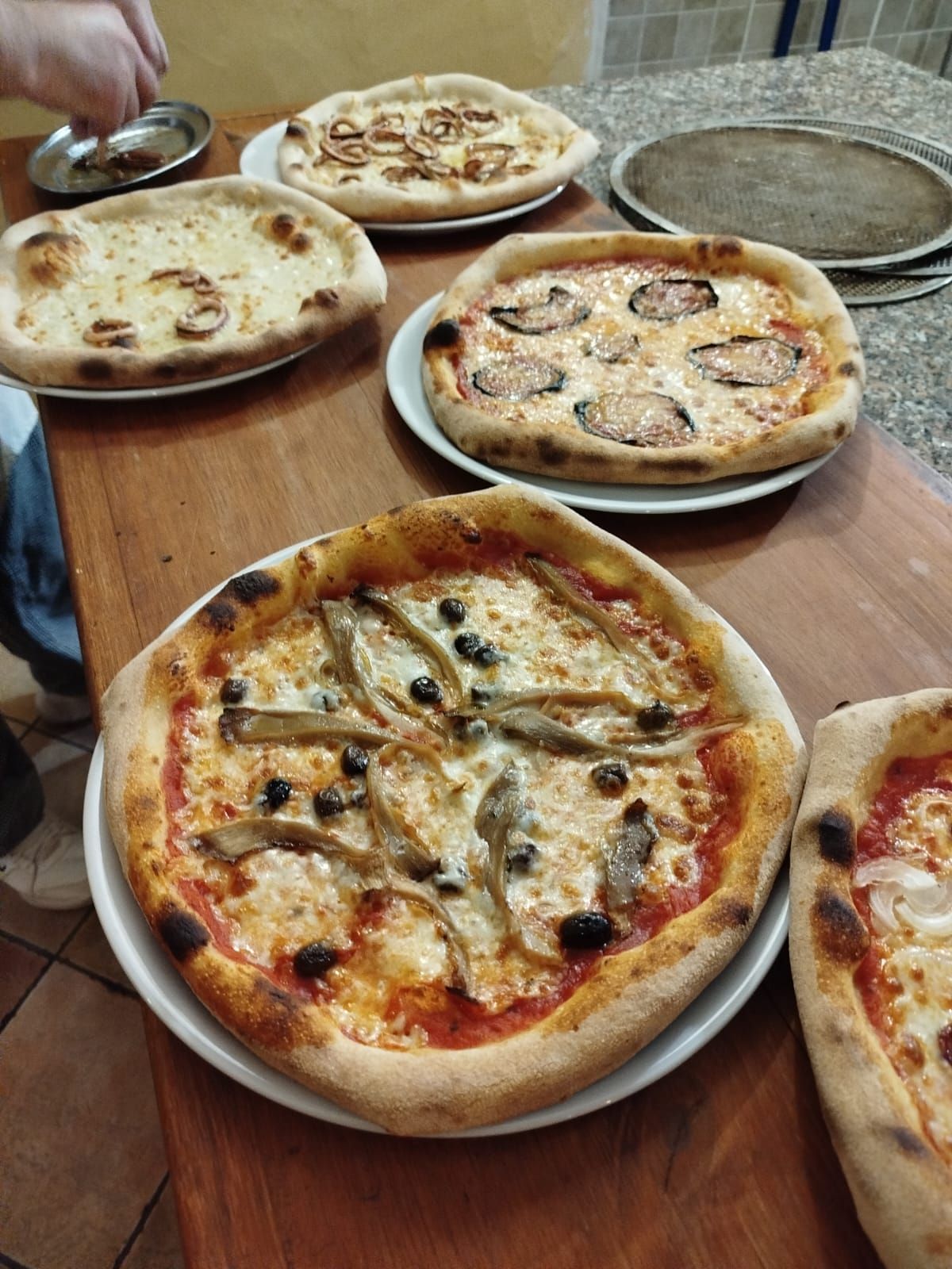 Su un tavolo di legno si trovano diverse pizze, tra cui una margherita e una ai funghi, servite su piatti bianchi.