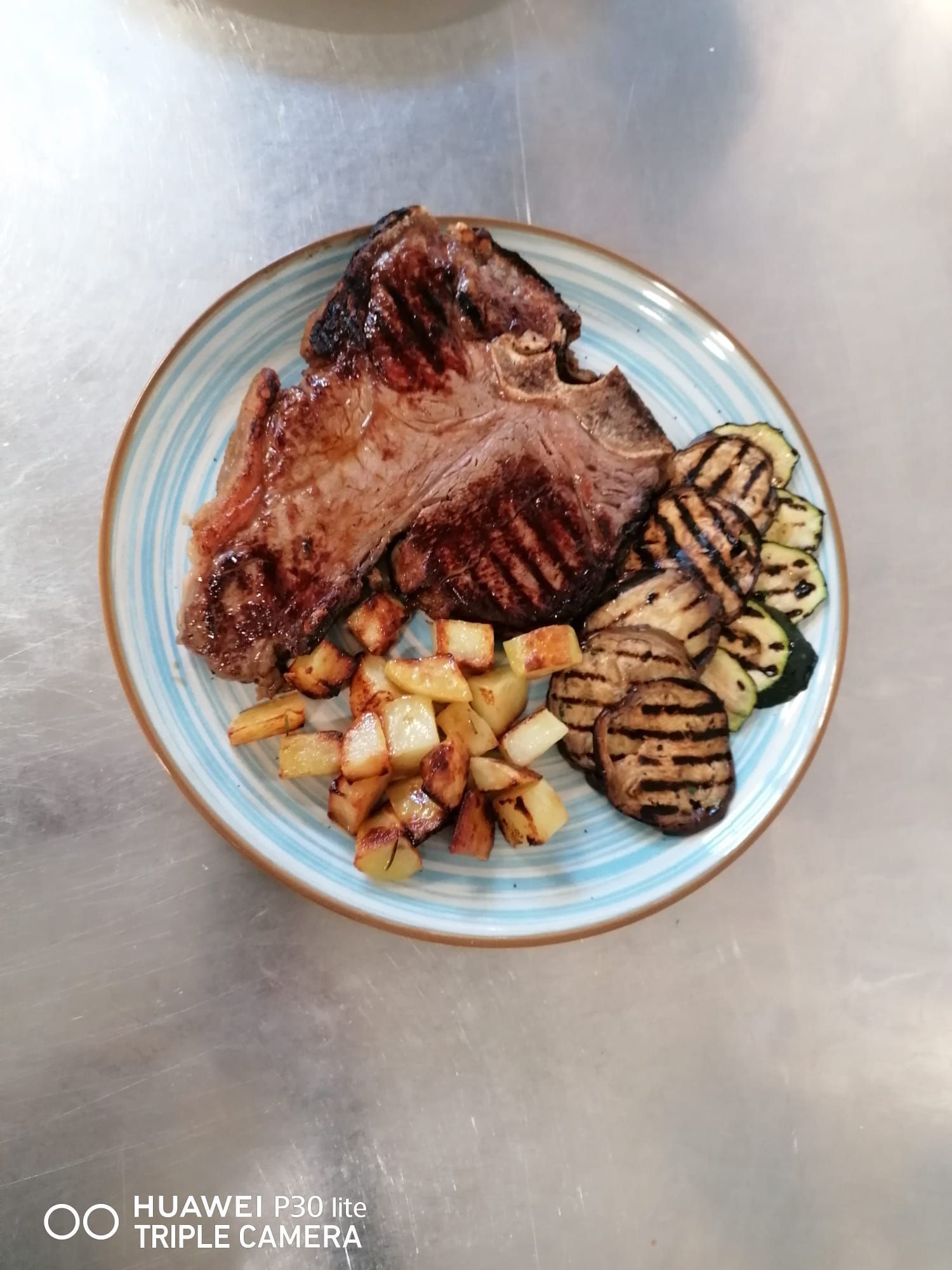 Carne alla griglia con patate arrosto e verdure a fette su un piatto azzurro chiaro.