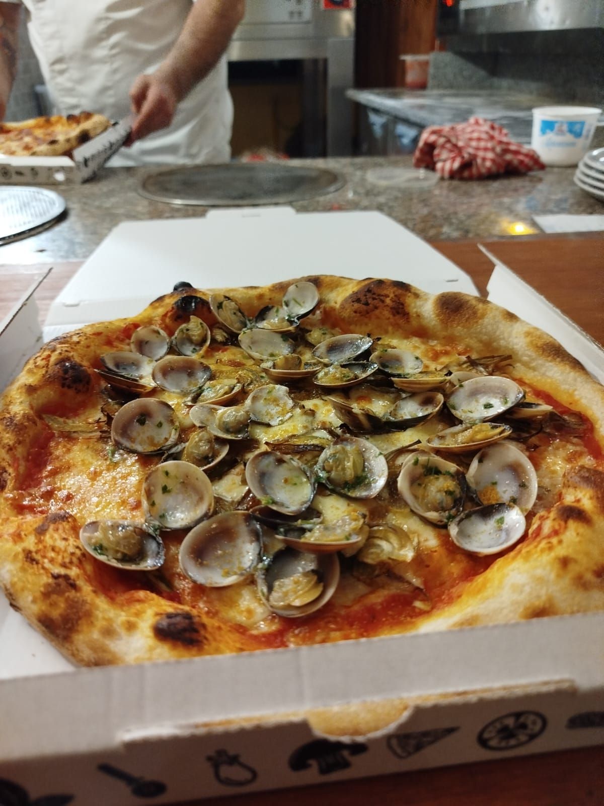 Pizza alle vongole in una scatola bianca su un bancone, con un cuoco visibile sullo sfondo.