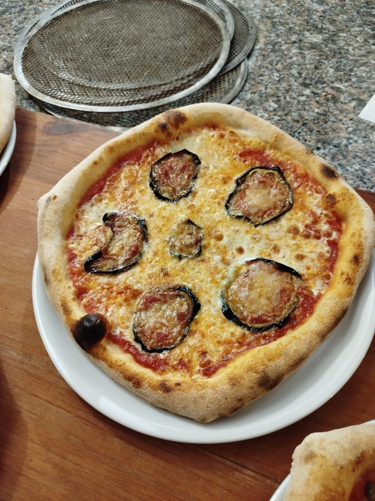 Pizza quadrata al formaggio con fette di melanzane su un piatto bianco appoggiato su un tagliere di legno.