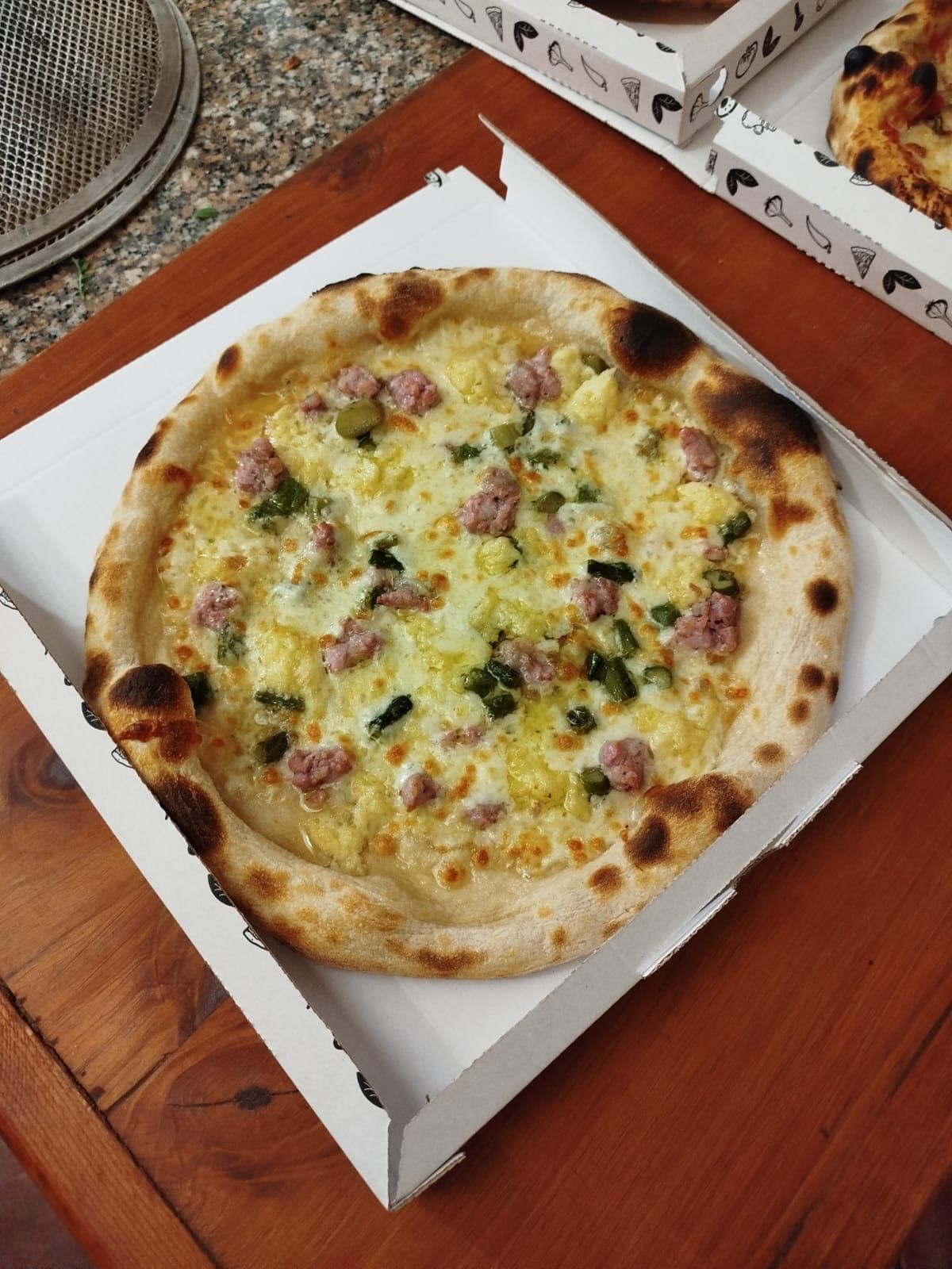 Pizza in una scatola bianca aperta su un tavolo di legno, condita con formaggio ed erbe aromatiche.