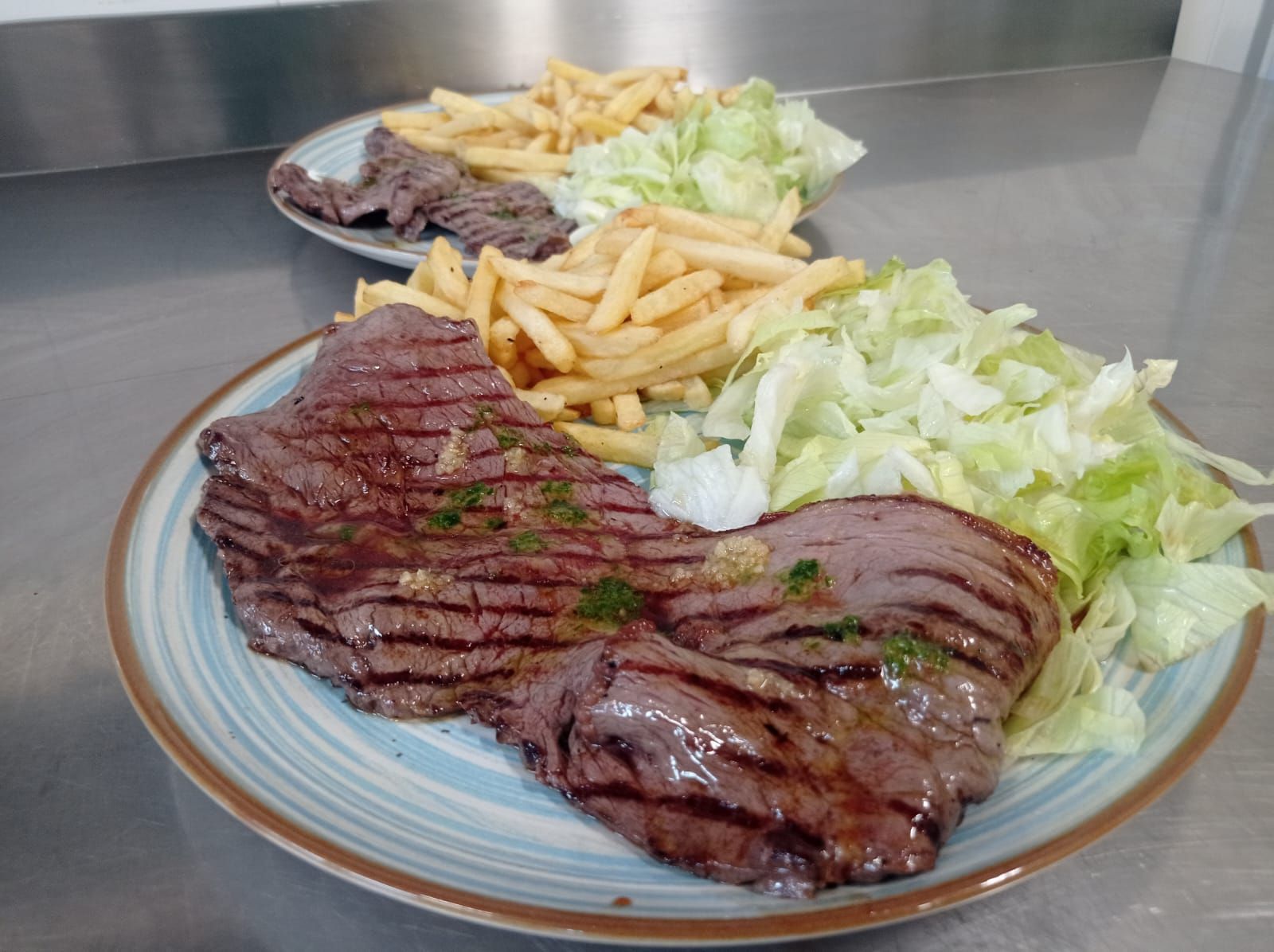 Piatti di bistecca alla griglia con patatine fritte e lattuga tritata su un bancone in acciaio inossidabile.