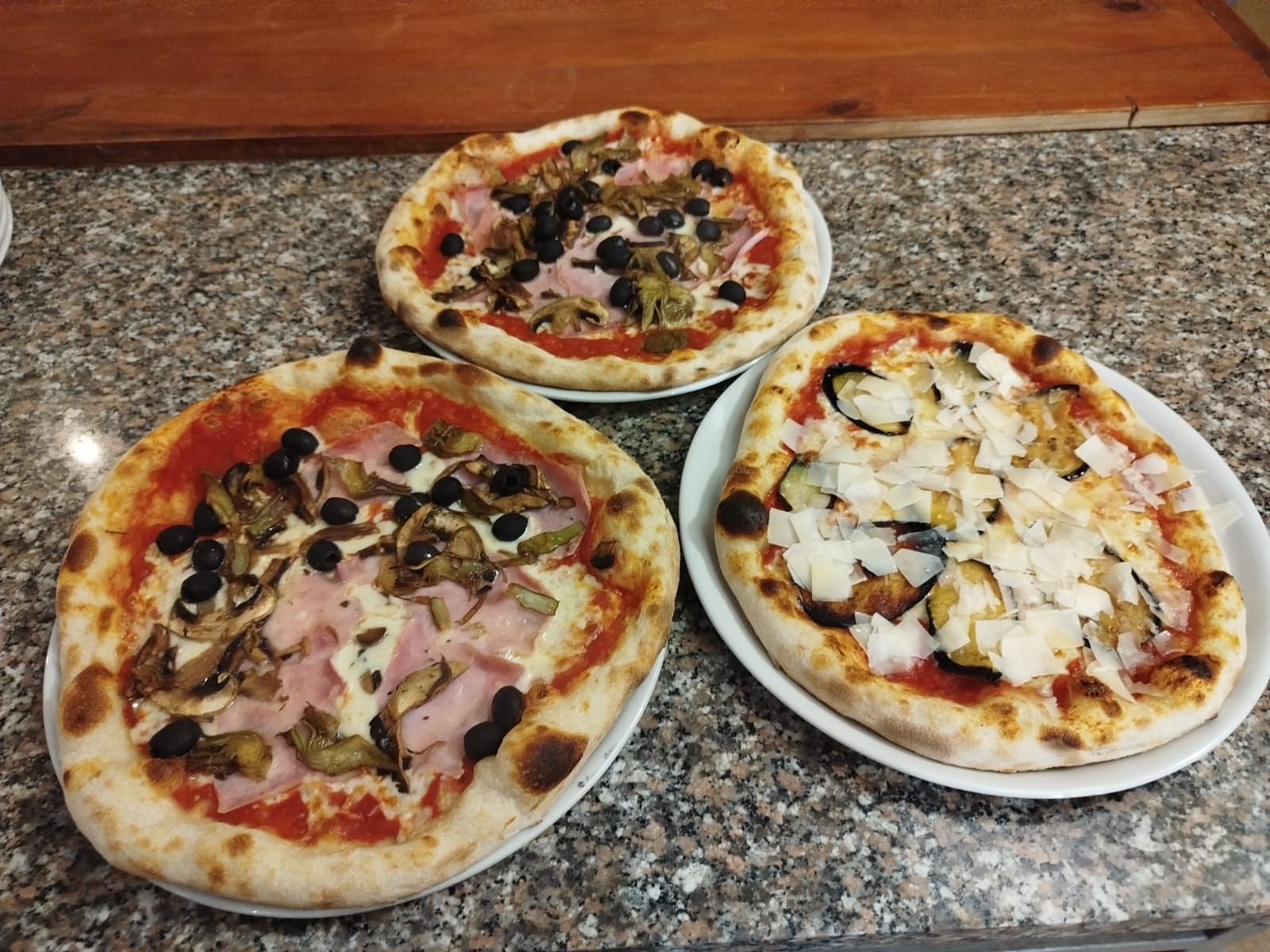 Tre pizze su un bancone di granito, condite con olive, prosciutto, funghi e cipolle.