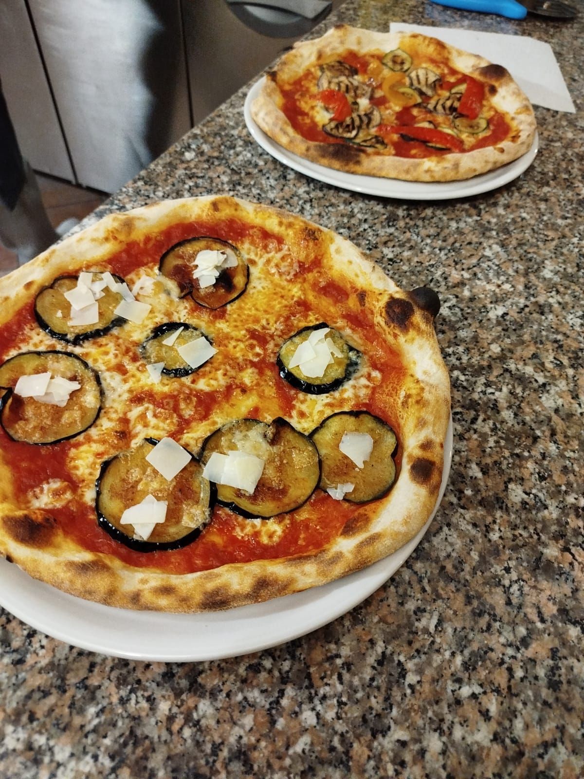 Due pizze fatte in casa su un piano di lavoro in granito, una con melanzane e formaggio, l'altra con pomodori e basilico.