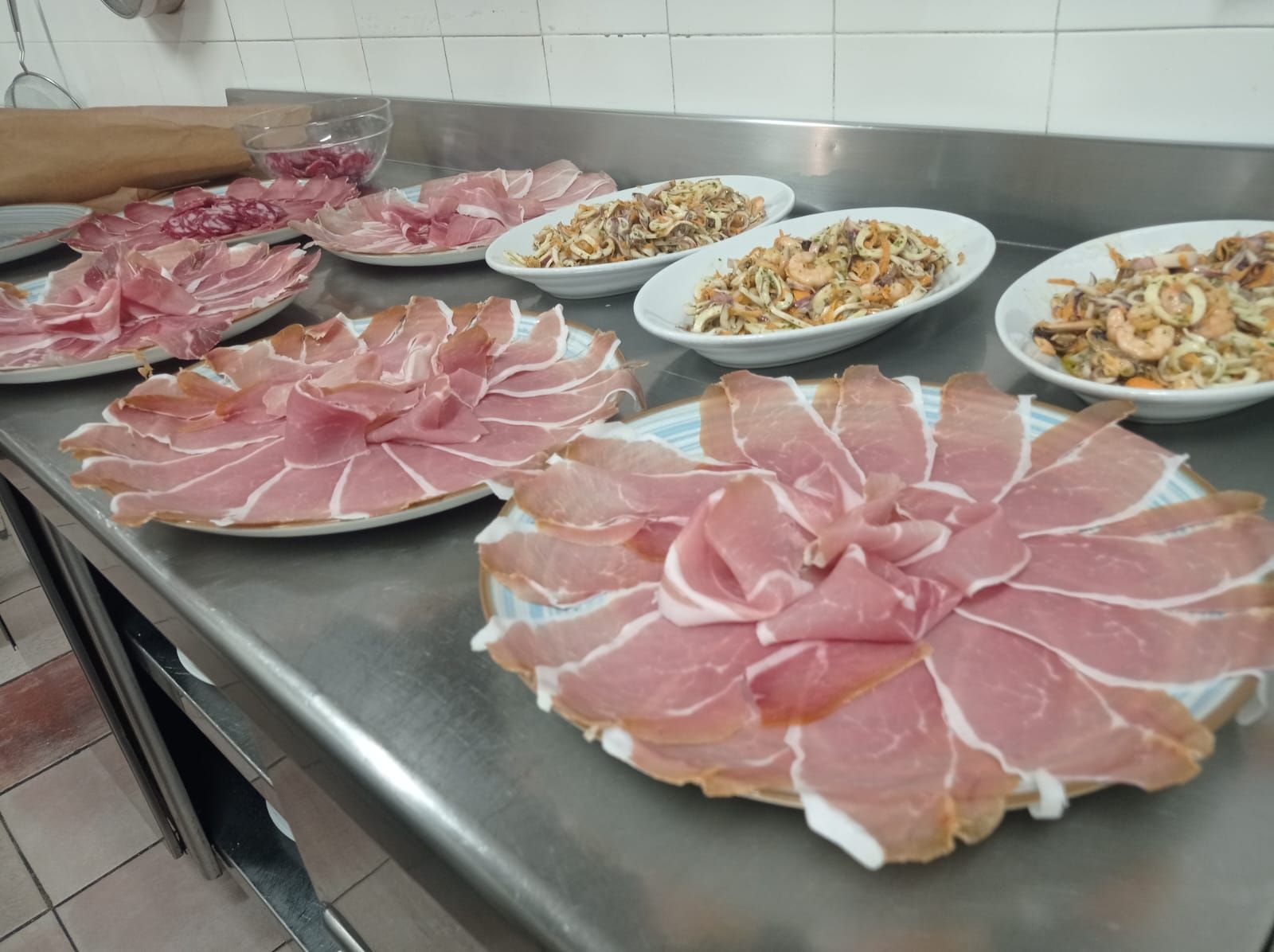 Piatti di prosciutto crudo affettato e antipasti di pesce su un bancone da cucina in acciaio inossidabile