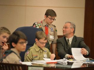 A group of boy scouts are sitting at a table with a man in a suit