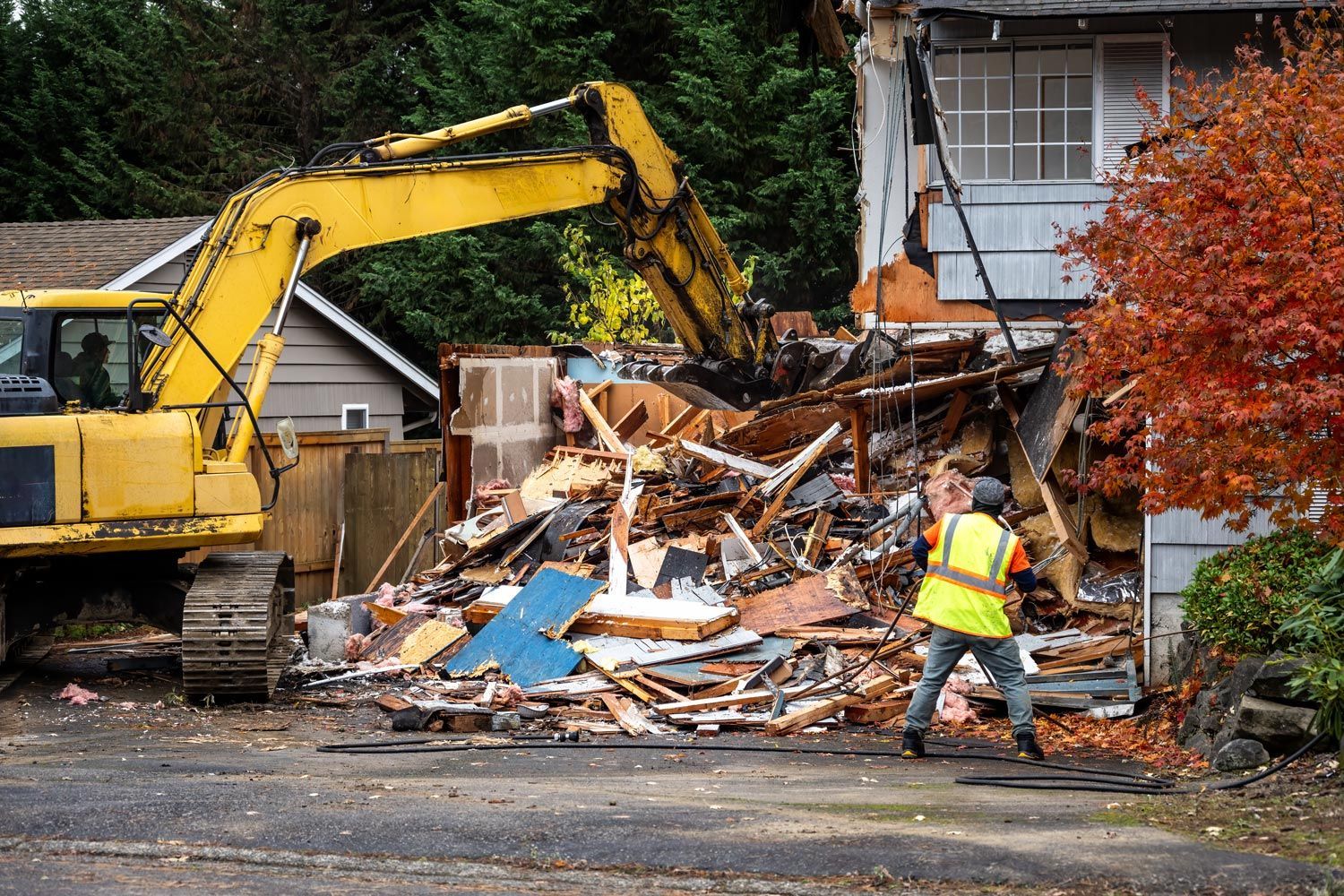 Yellow excavator demolishing a house as a worker in a reflective vest stands beside rubble Yellow excavator demolishing a house as a worker in a reflective vest stands beside rubble