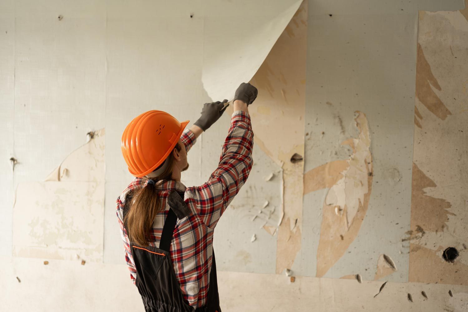 Person in orange hard hat scrapes peeling paint from a wall with a putty knife. Person in orange hard hat scrapes peeling paint from a wall with a putty knife.