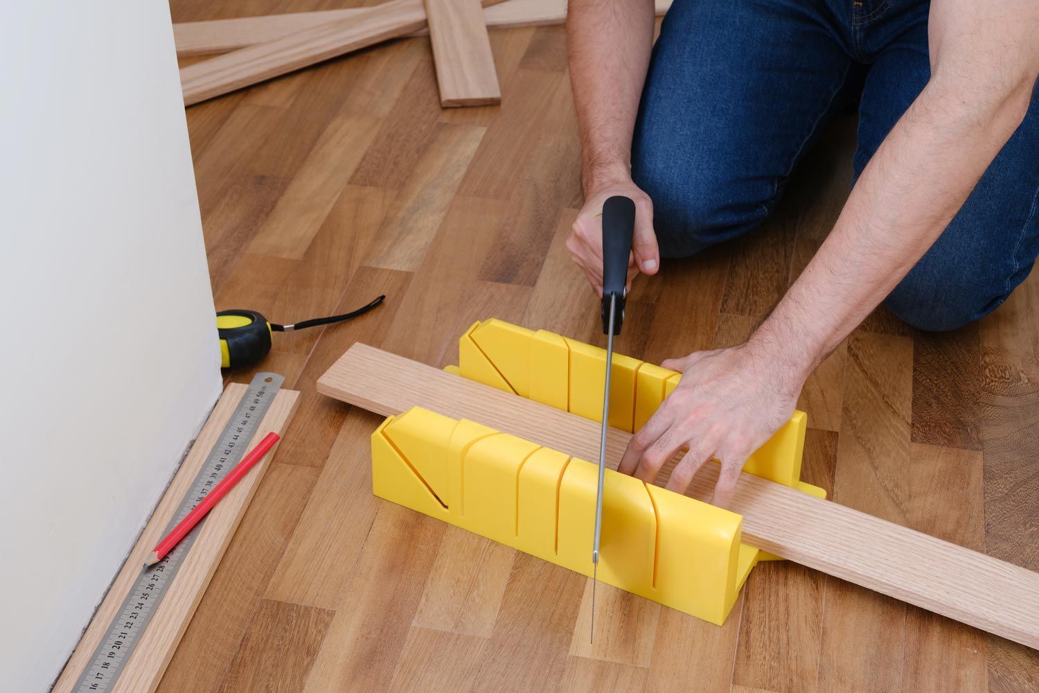 Person cutting a wooden board on the floor with a handsaw, surrounded by yellow clamps and measuring tools Person cutting a wooden board on the floor with a handsaw, surrounded by yellow clamps and measuring tools