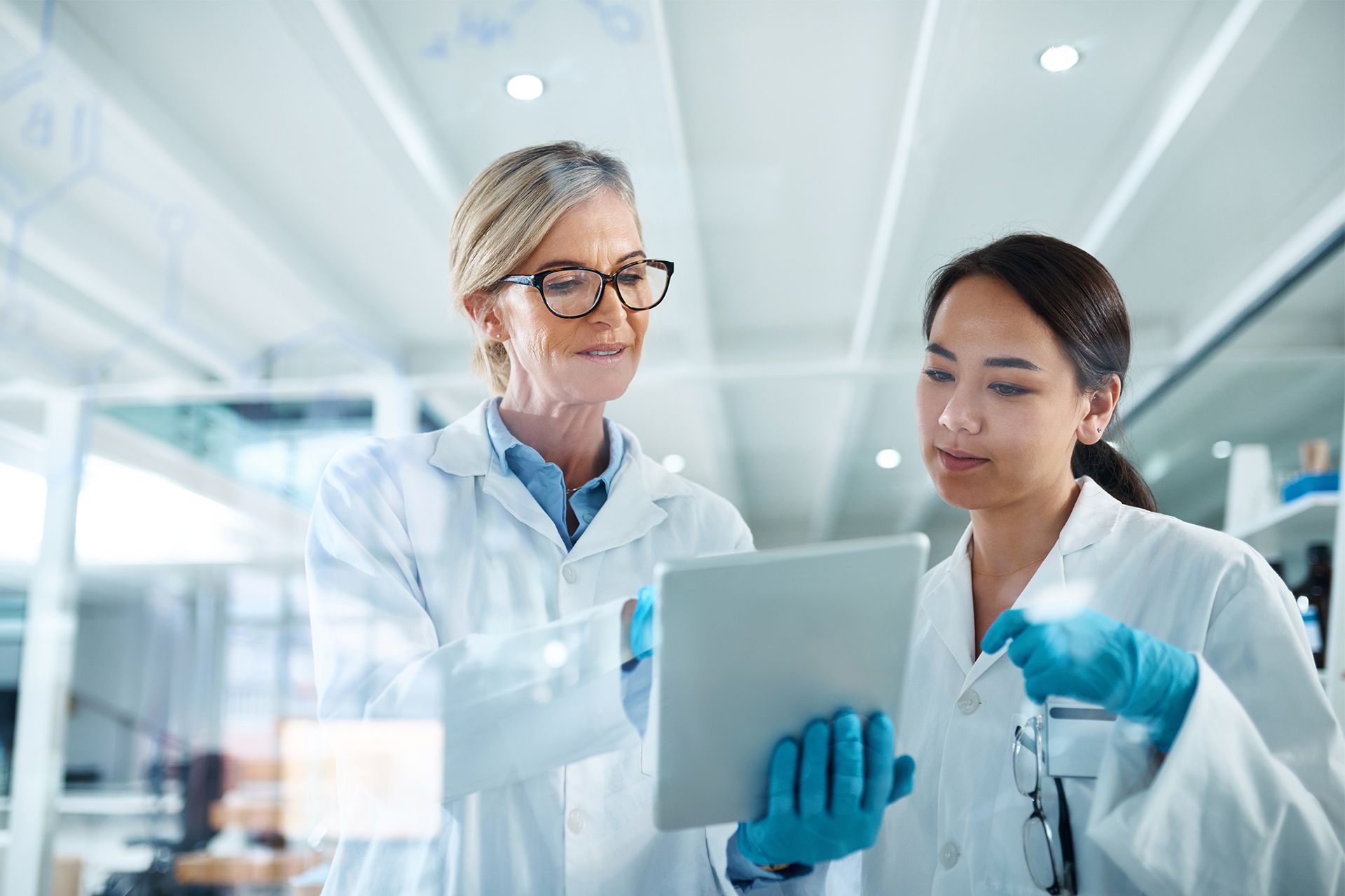 Two scientists in lab coats and blue gloves reviewing a tablet in a bright laboratory