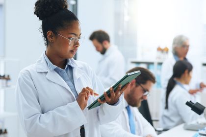 Scientist in a white lab coat using a tablet in a bright lab, with coworkers blurred in the background.