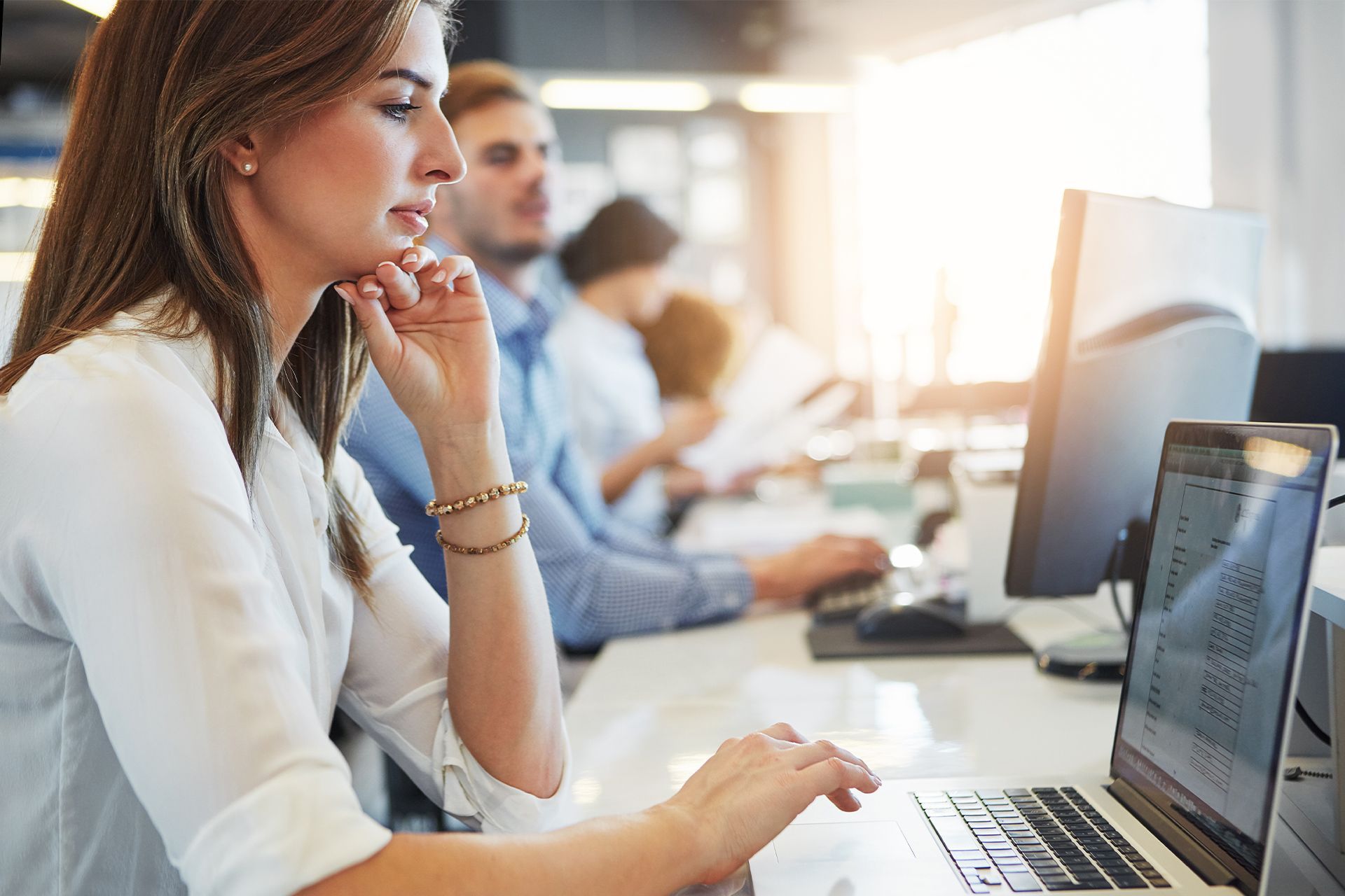 Woman working at a computer in a bright office, with colleagues seated behind her.
