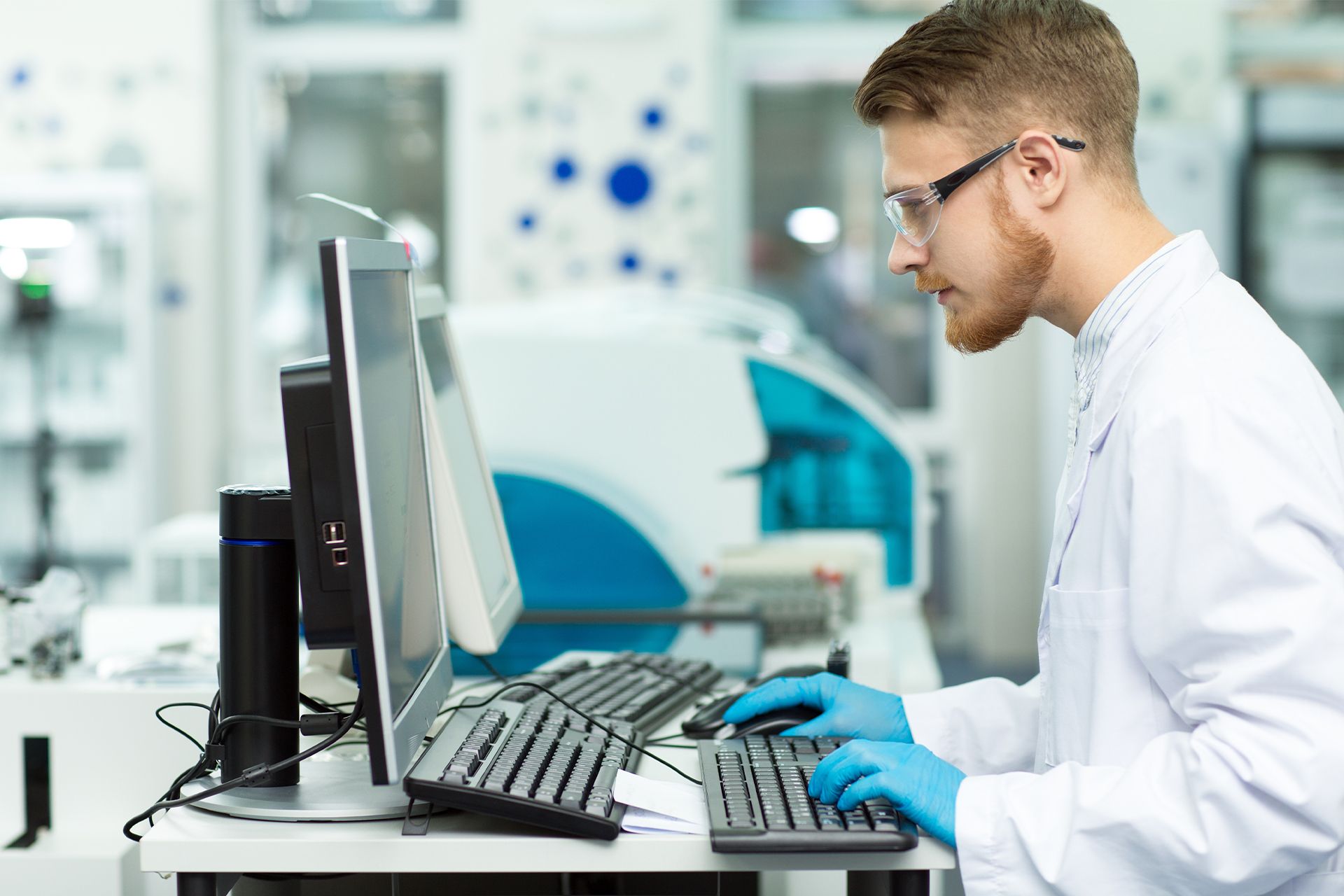Scientist typing at a lab computer while wearing a white coat and blue gloves in a modern laboratory