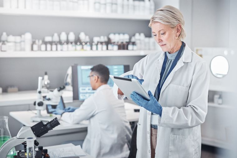 Scientists in white coats working in a bright laboratory, one using a microscope and another reading a tablet