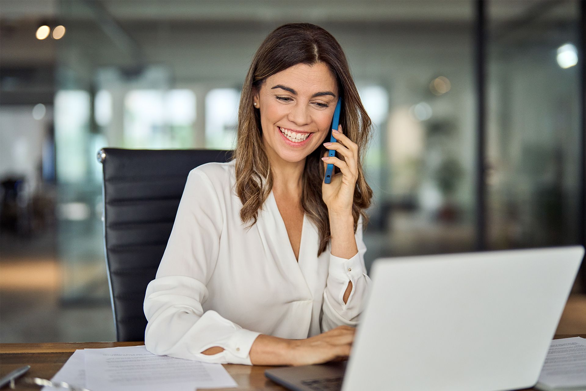 Woman smiling while talking on a phone at a laptop in an office