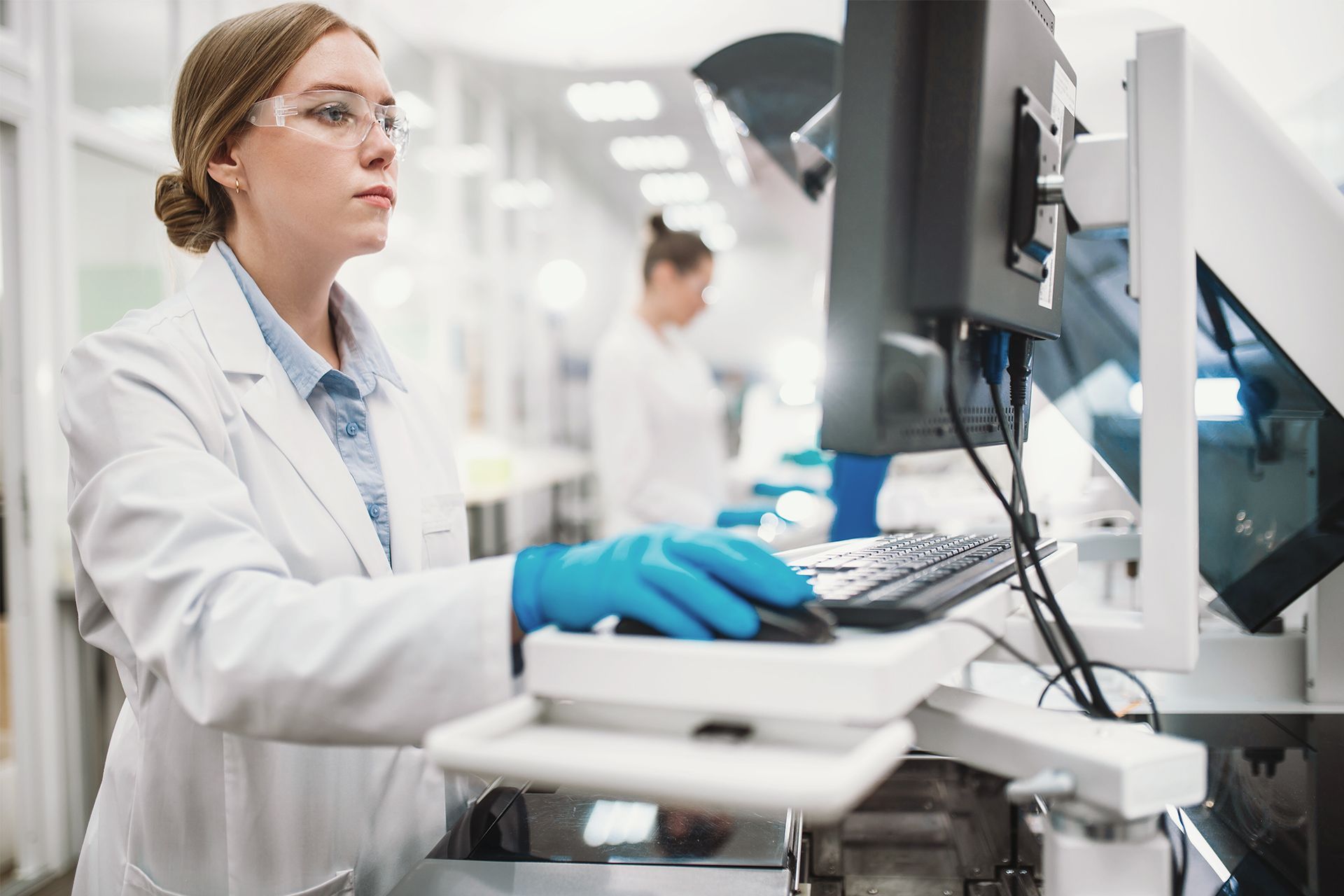Lab technician in a white coat and blue gloves working at a computer in a clinical lab.