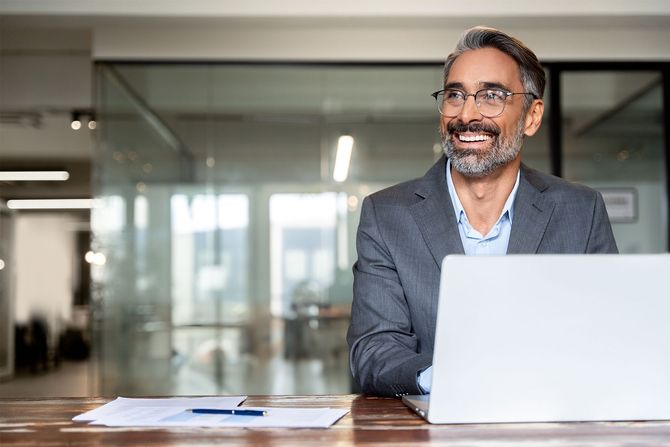 Smiling man in a blazer working on a laptop at a desk in a modern office