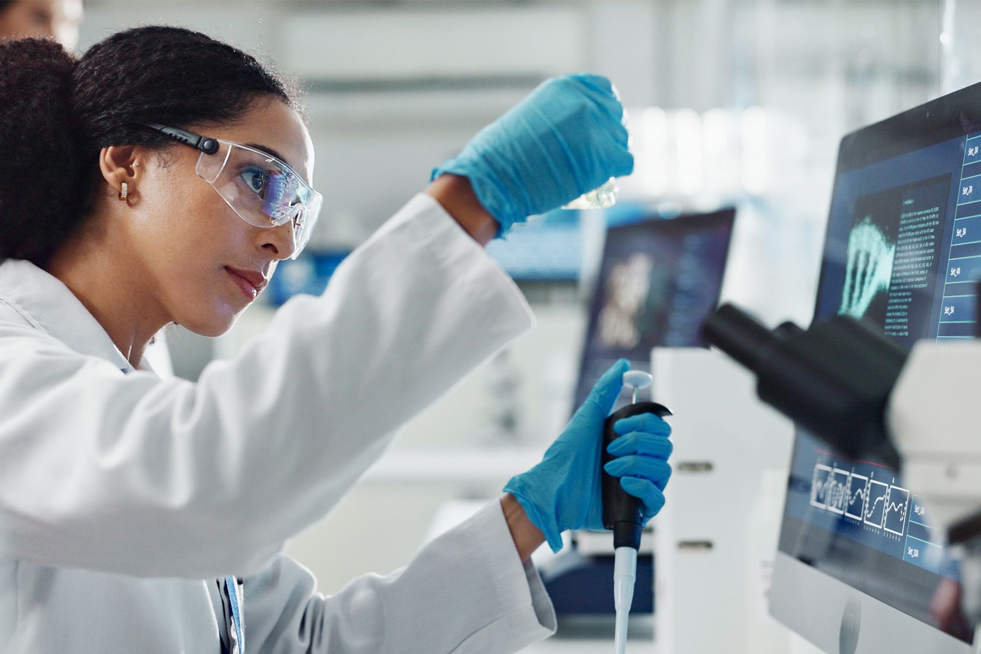 Scientist in lab coat and gloves using a pipette beside a computer monitor in a laboratory