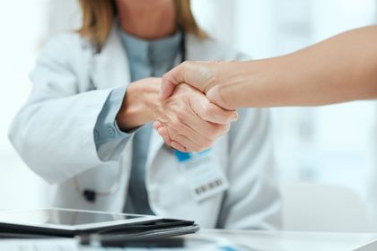 Handshake between a doctor and patient in a clinic, with a stethoscope and clipboard on the desk.