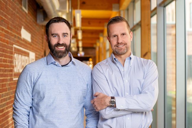 Two men stand in a hallway, one in a blue shirt and one in a white checkered shirt.