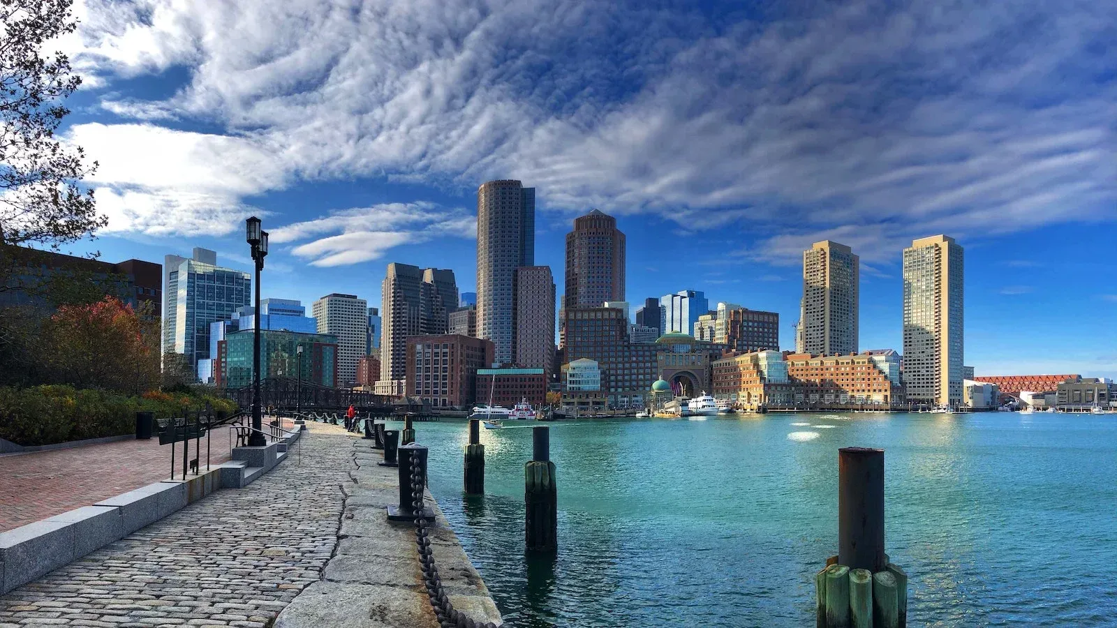 Waterfront city skyline with tall buildings, blue water, and a stone promenade under a cloudy sky
