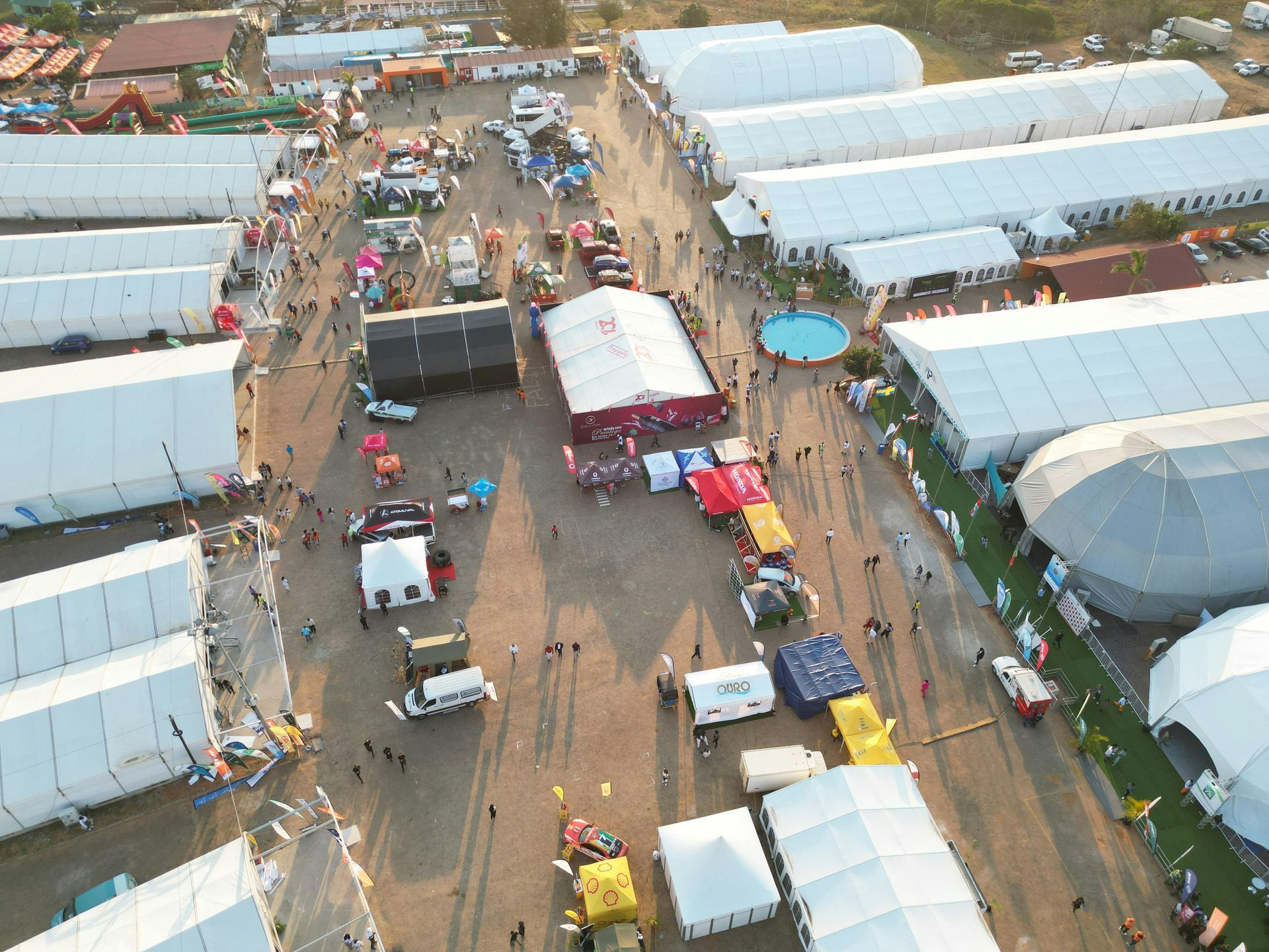 Aerial view of a crowded outdoor market with white tents, vehicles, and many people on dusty paths.