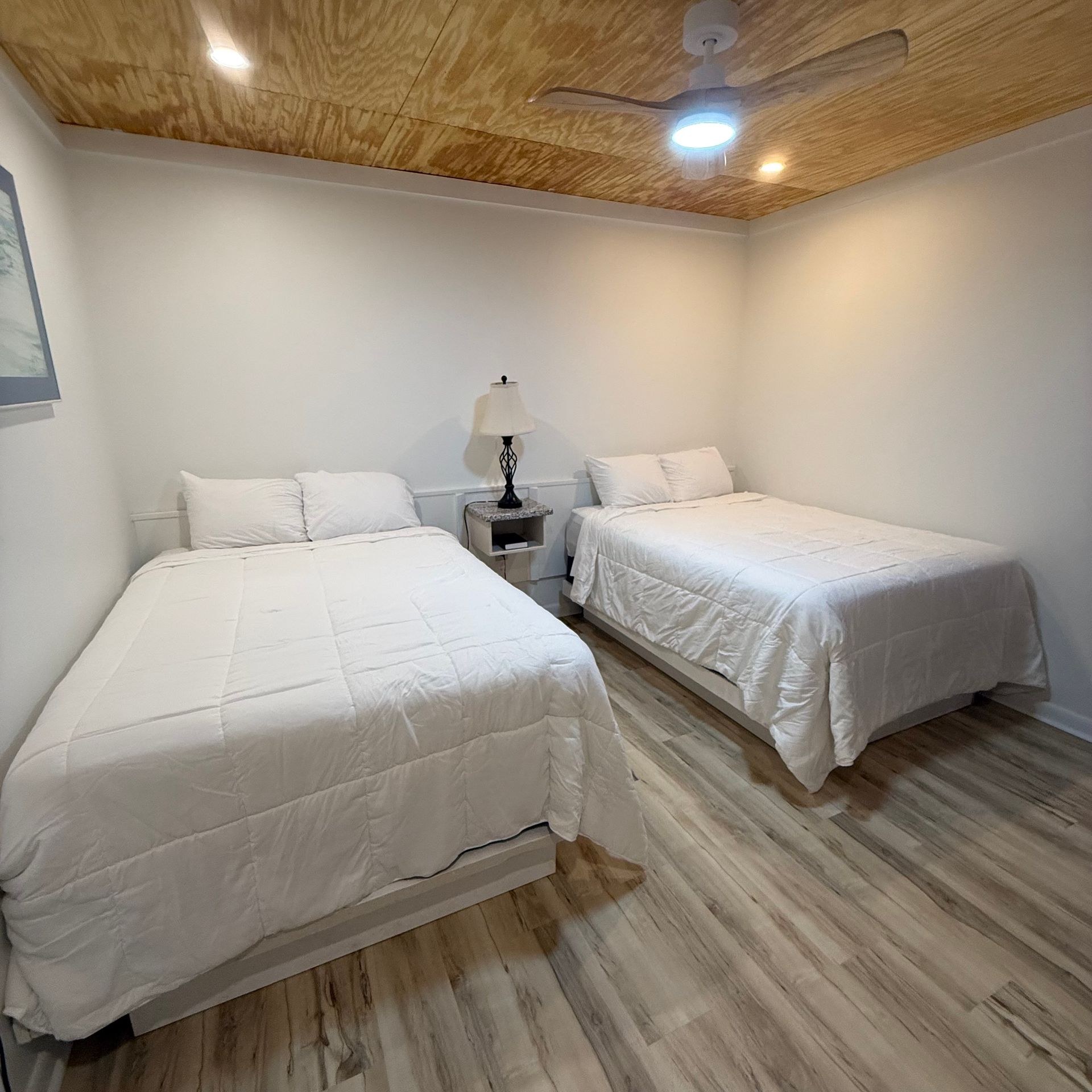 Small hotel room with two beds, a window air conditioner, and a nightstand with a lamp and framed art.