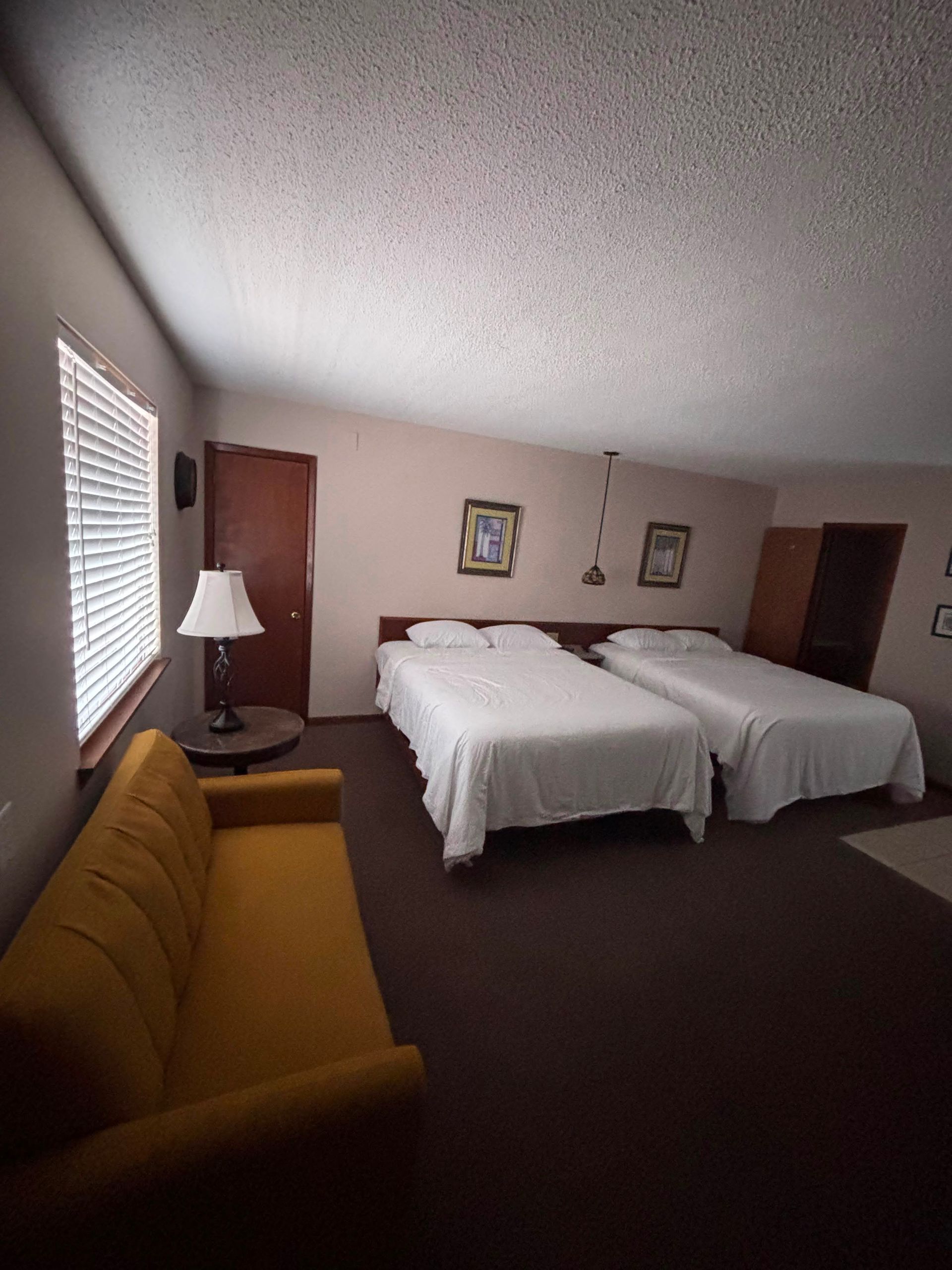 Motel room with two double beds, mustard sofa, window blinds, and a lamp on a side table.
