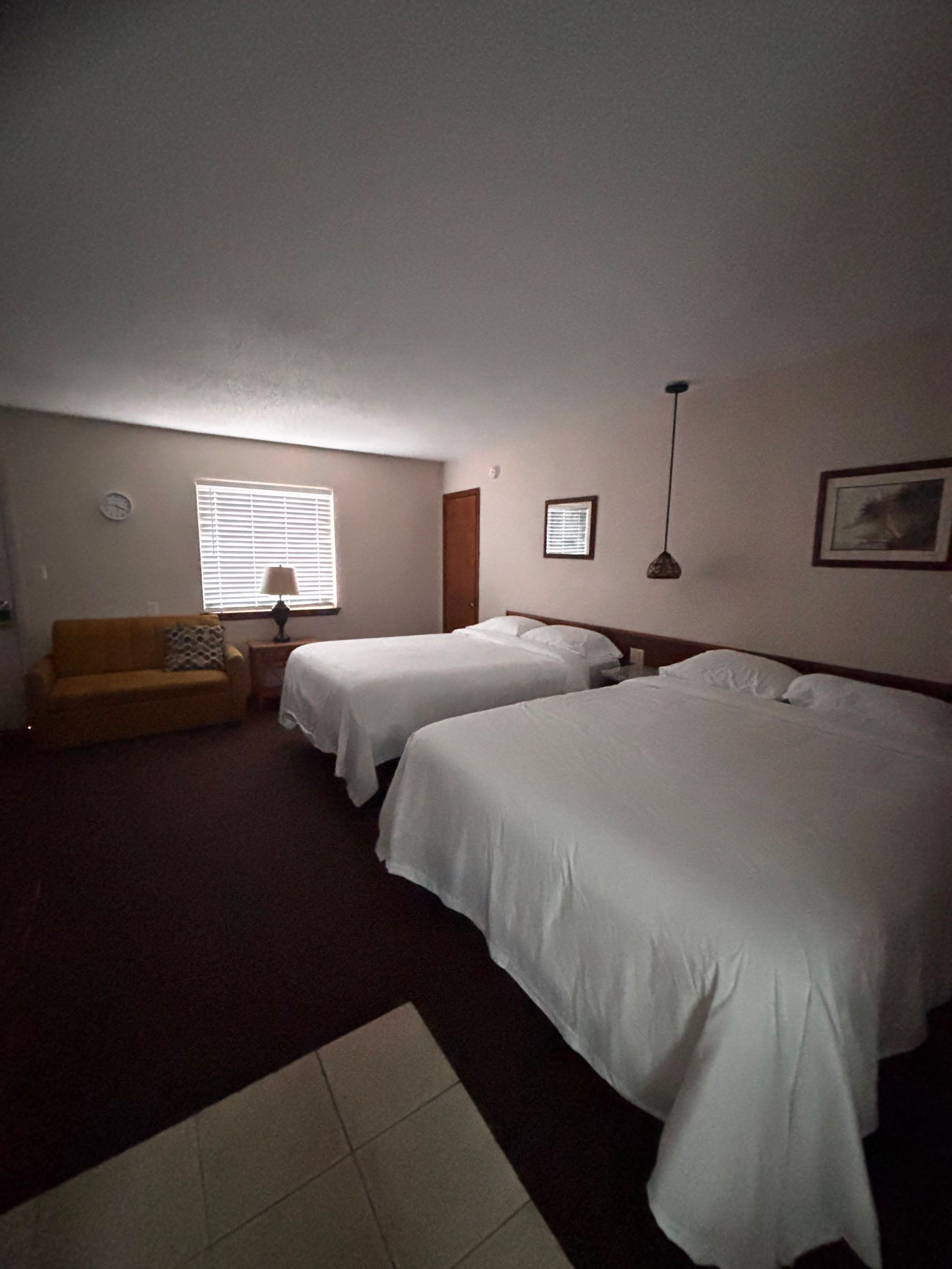 Hotel room with two white-covered beds, a window, lamp, desk, and framed wall art.