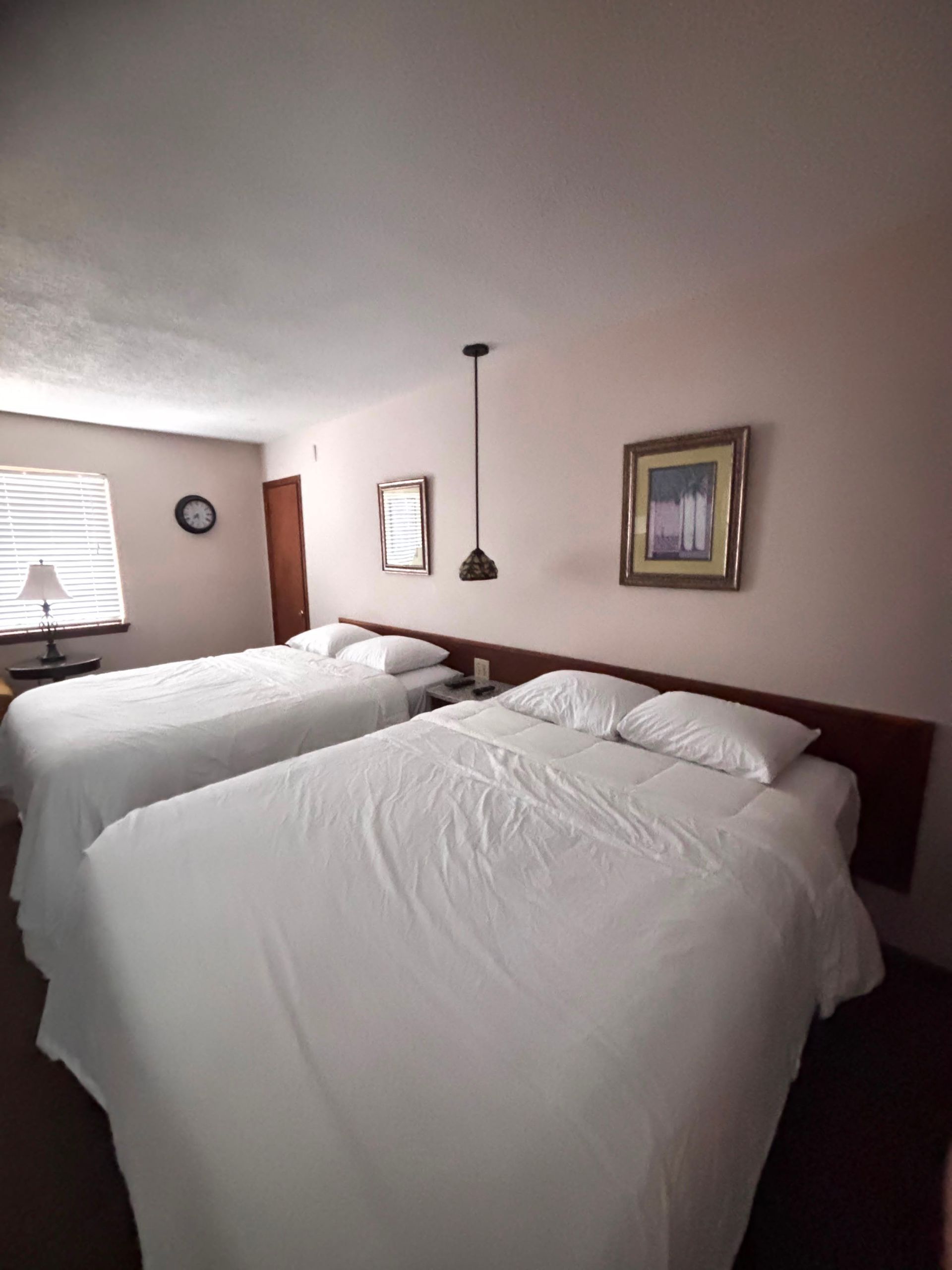 Hotel room with two unmade beds, beige walls, and a window with blinds and a small table.