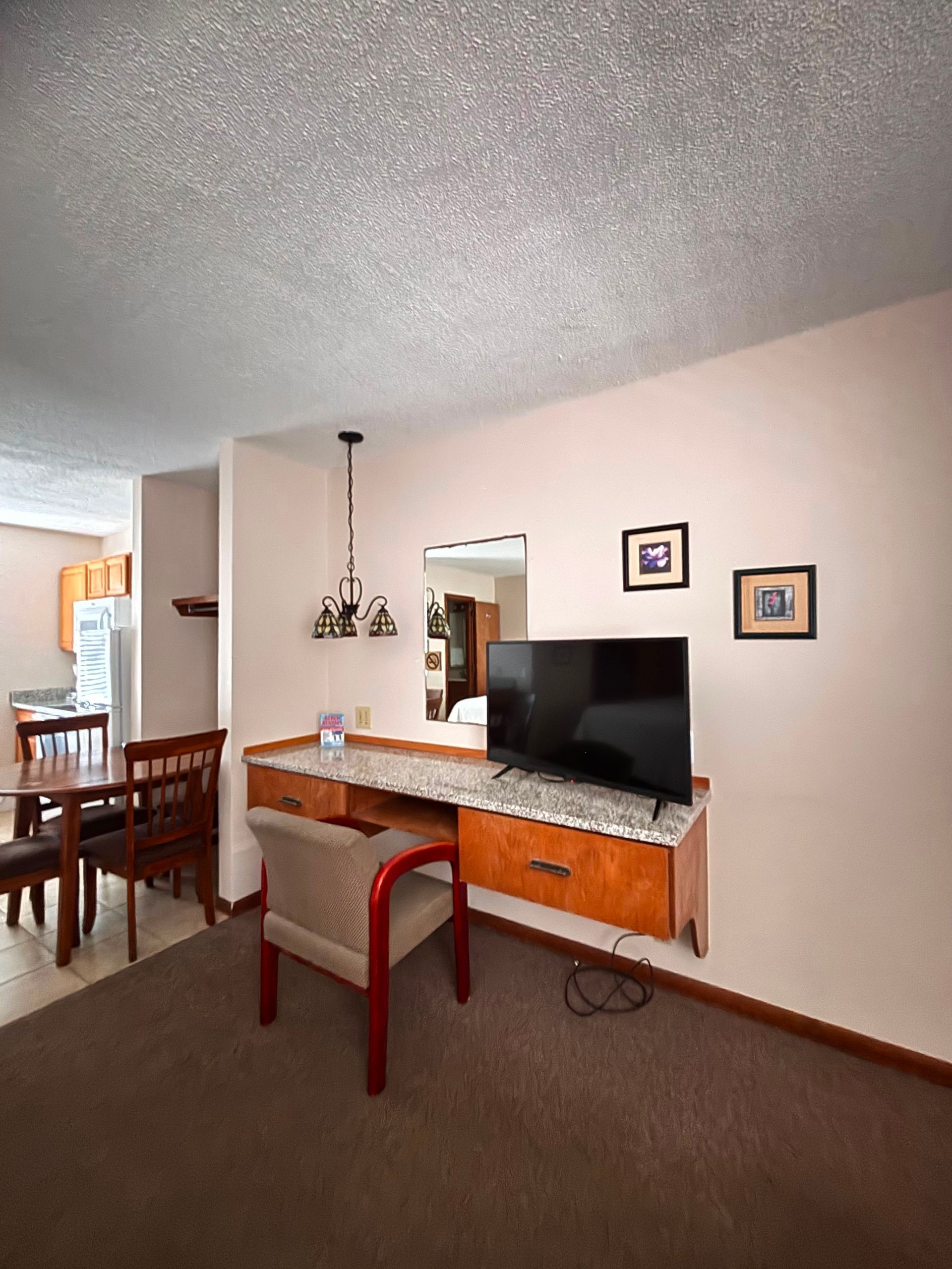 Home office nook with wooden desk, computer monitor, red chair, and dining area in the background