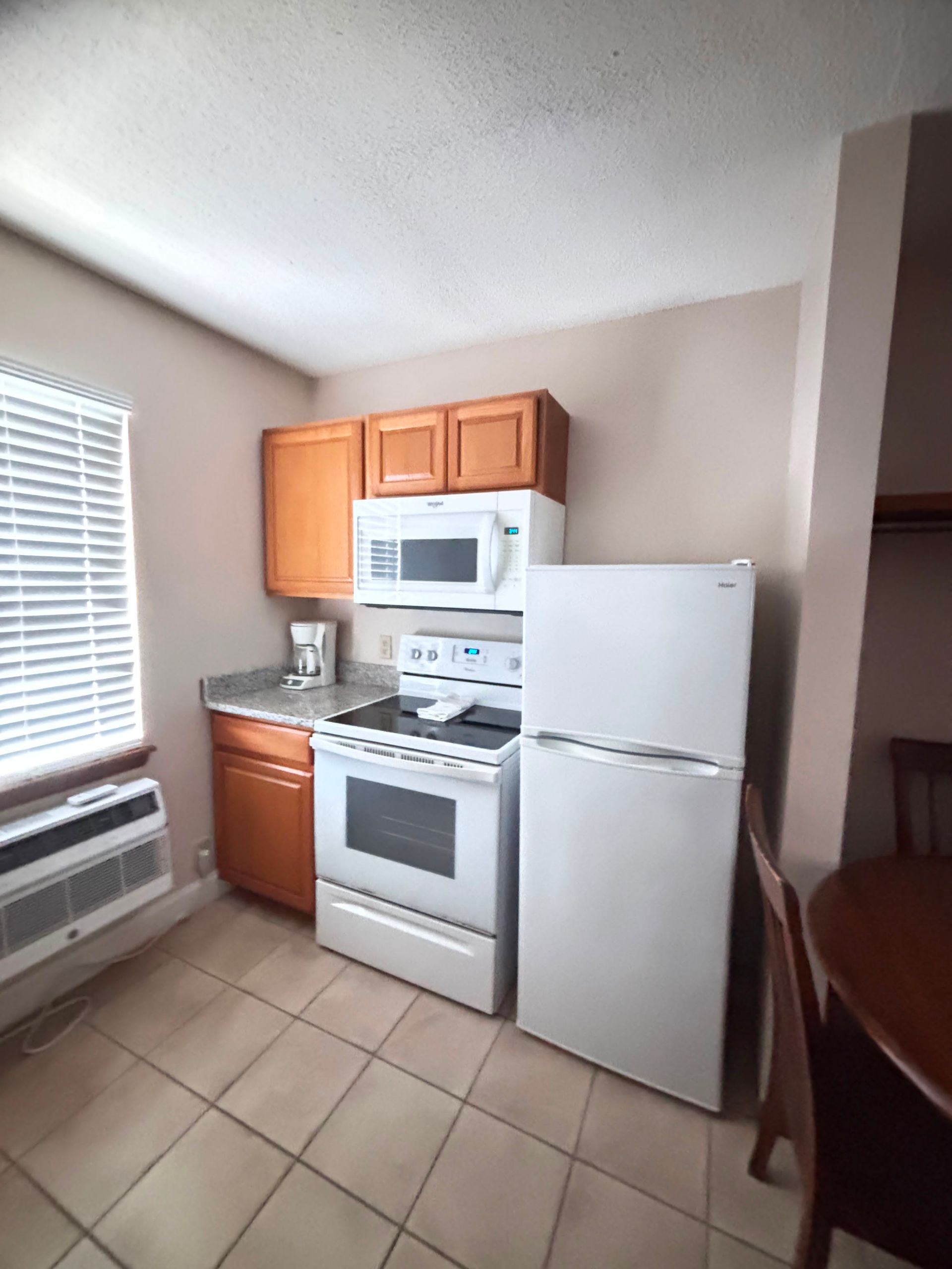 Small kitchen with white appliances, wood cabinets, tile floor, and a window with blinds.