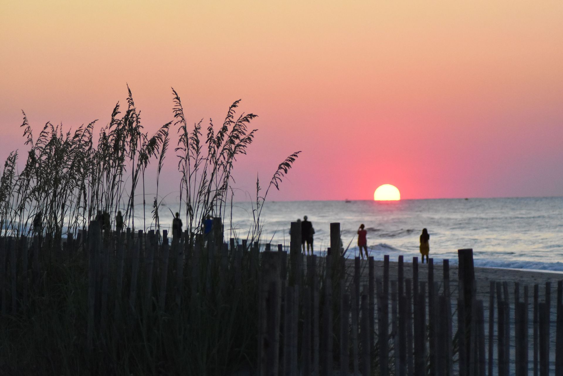 Sunset over a beach with silhouetted dunes and people standing by the waterline