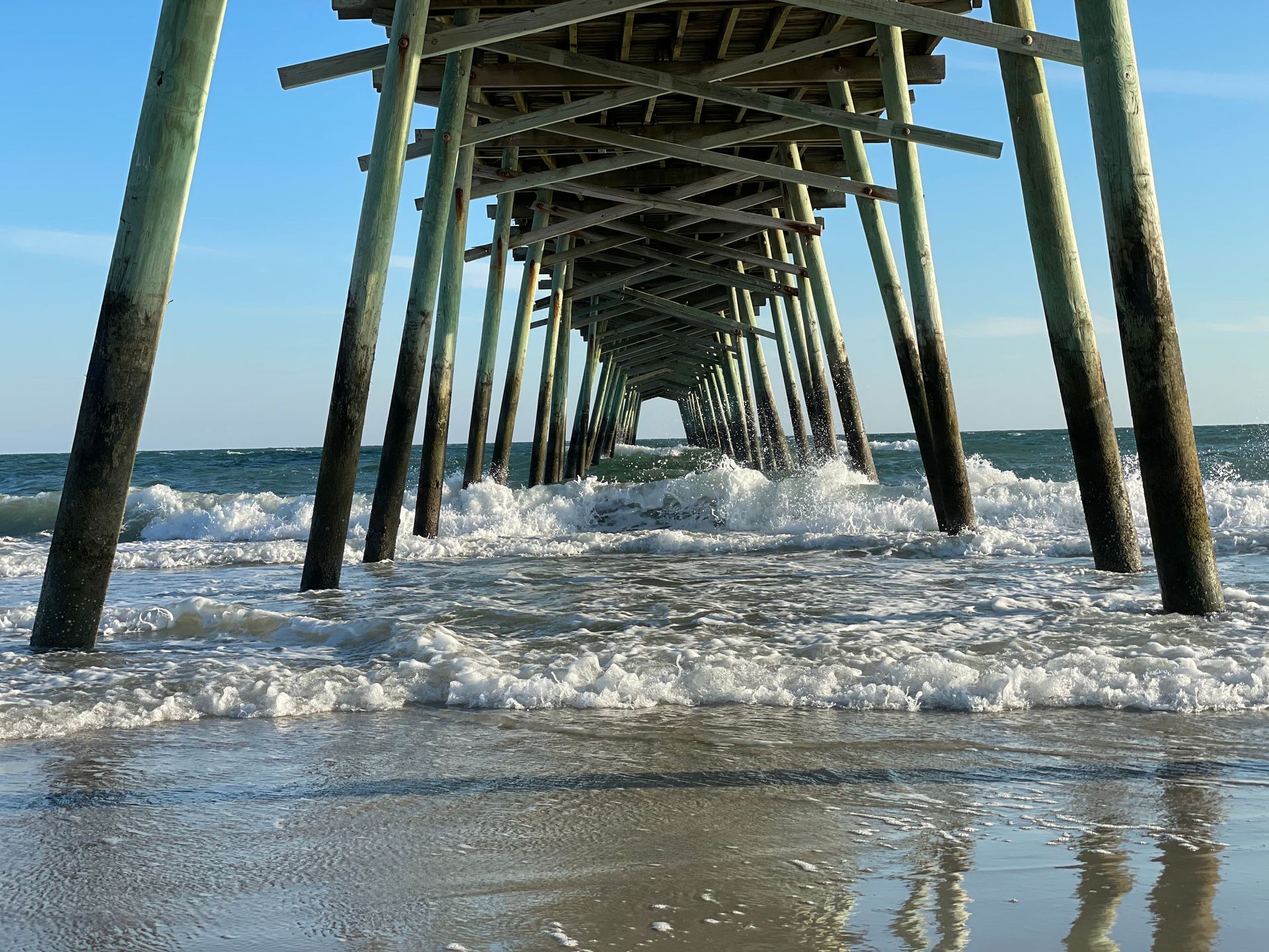 View from beneath a pier with waves crashing around its wooden pilings above a sandy beach